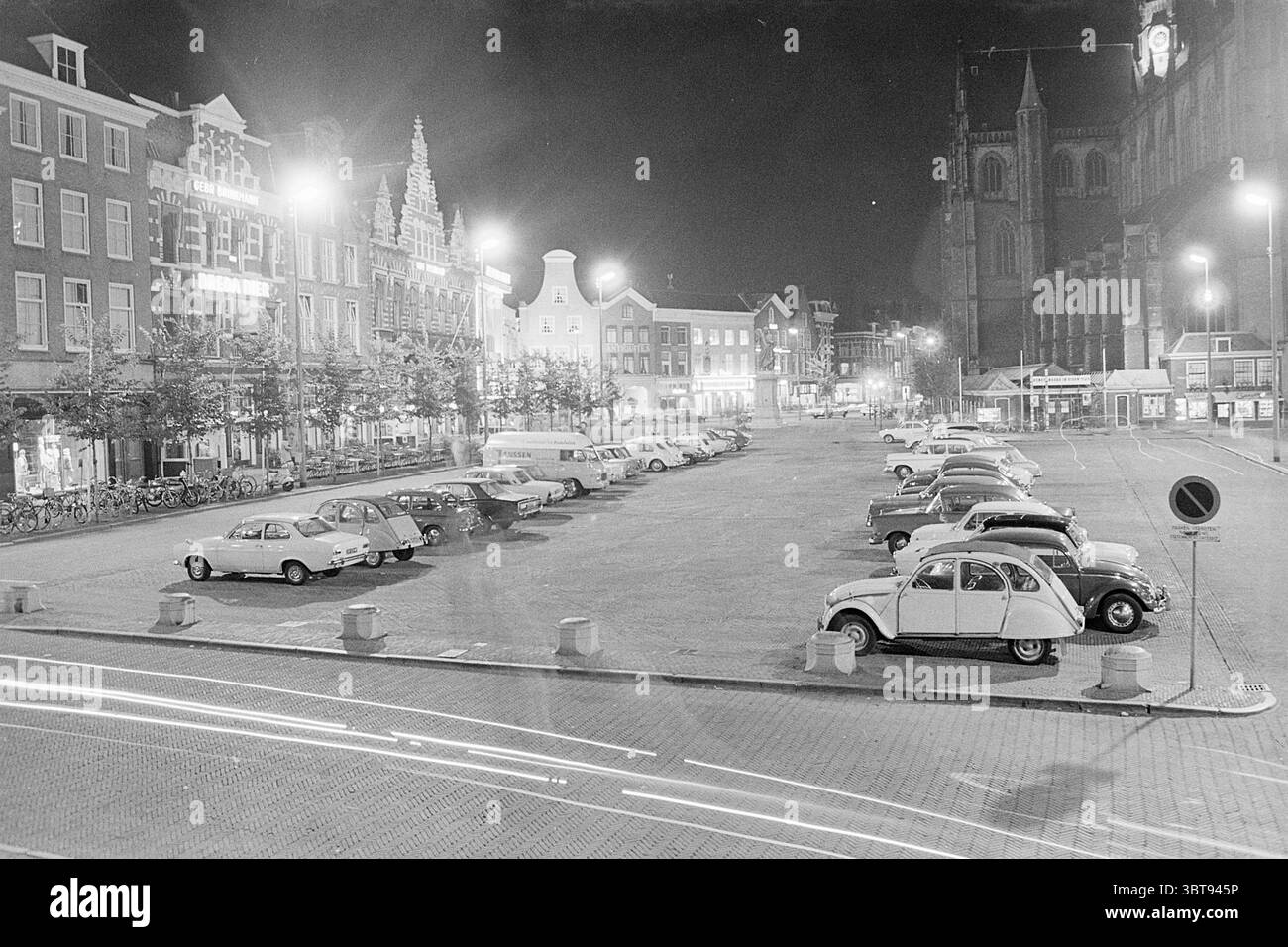 Auto su Grote Markt Grote Markt Haarlem Grote Markt Paesi Bassi, Whizgle News, Dutch Desk, Paesi Bassi, 1950 - 2000 il 20-09-1969. L'immagine mostra questi argomenti. La scena è un paesaggio urbano notturno, caratterizzato da un ampio parcheggio per lo più vuoto, con alcune auto d'epoca ben allineate. Il layout è caratterizzato da un mix di modelli classici, che mostrano contorni arrotondati e design distintivi di un'epoca passata, i loro colori vanno dai bianchi e grigi tenui alle tonalità più profonde, il tutto leggermente attenuato sotto l'oscurità della notte. Nei dintorni del parcheggio si trova un affascinante edificio storico Foto Stock