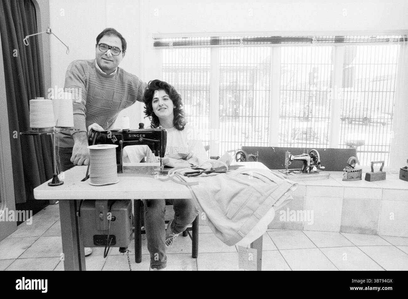 Mario Tailor Kleine Houtstraat Clothing Haarlem Kleine Houtstraat Nederland, Whizgle News, Dutch Desk, Paesi Bassi, 1950 - 2000 il 02-04-1986. Questi sono gli argomenti nell'immagine. In uno spazio luminoso e ben illuminato, due persone occupano un tavolo centrale in legno adornato con varie forniture da cucire. Il tavolo presenta rocchetti di filo in tenui colori pastello, a indicare un ambiente incentrato sull'artigianato. Una classica macchina da cucire, posizionata in modo preminente, enfatizza un senso di arte e abilità. La parete posteriore è rivestita da grandi finestre, che consentono alla luce naturale di inondare la scena e proiettare ombre delicate acr Foto Stock