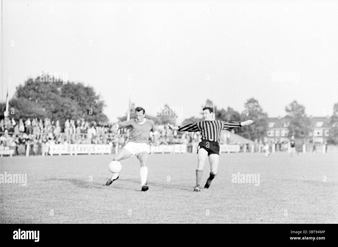 EDO - RCH Football EDO, Whizgle News, Dutch Desk, Paesi Bassi, 1950 - 2000 il 06-08-1967. L'immagine mostra questi argomenti. La scena cattura un momento di energia su un campo da calcio erboso. In primo piano, due giocatori sono impegnati in un gioco competitivo. Uno, vestito con una vivace maglia rossa e pantaloncini bianchi, controlla sapientemente la palla con il piede. La sua espressione è focalizzata, evidenziando la sua determinazione a mantenere il possesso. Il giocatore avversario, rivestito da una maglia bianca e nera a righe orizzontali con pantaloncini corti, si trova dietro di lui, tentando di sfidare la palla. Il backgrou Foto Stock