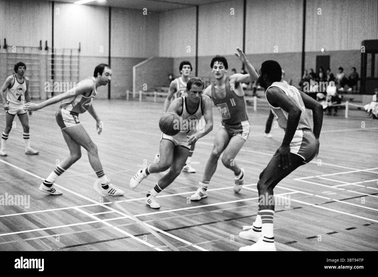 Partita di basket Akrides e 7UP., Whizgle News, Dutch Desk, Paesi Bassi, 1950 - 2000. Questi argomenti vengono visualizzati nell'immagine. La scena cattura un momento intenso in una partita di basket ambientata in una spaziosa palestra interna. Il pavimento in legno lucido si estende attraverso la cornice, riflettendo le luci superiori, che proiettano una luminosa illuminazione sul campo. Da un lato, un gruppo di giocatori, rivestiti di uniformi abbinate con numeri in grassetto, mostra una serie di espressioni, alcune mirate, altre determinate, evidenziando l'atmosfera competitiva. Sono impegnati in movimenti dinamici; un giocatore sta dribbling t Foto Stock