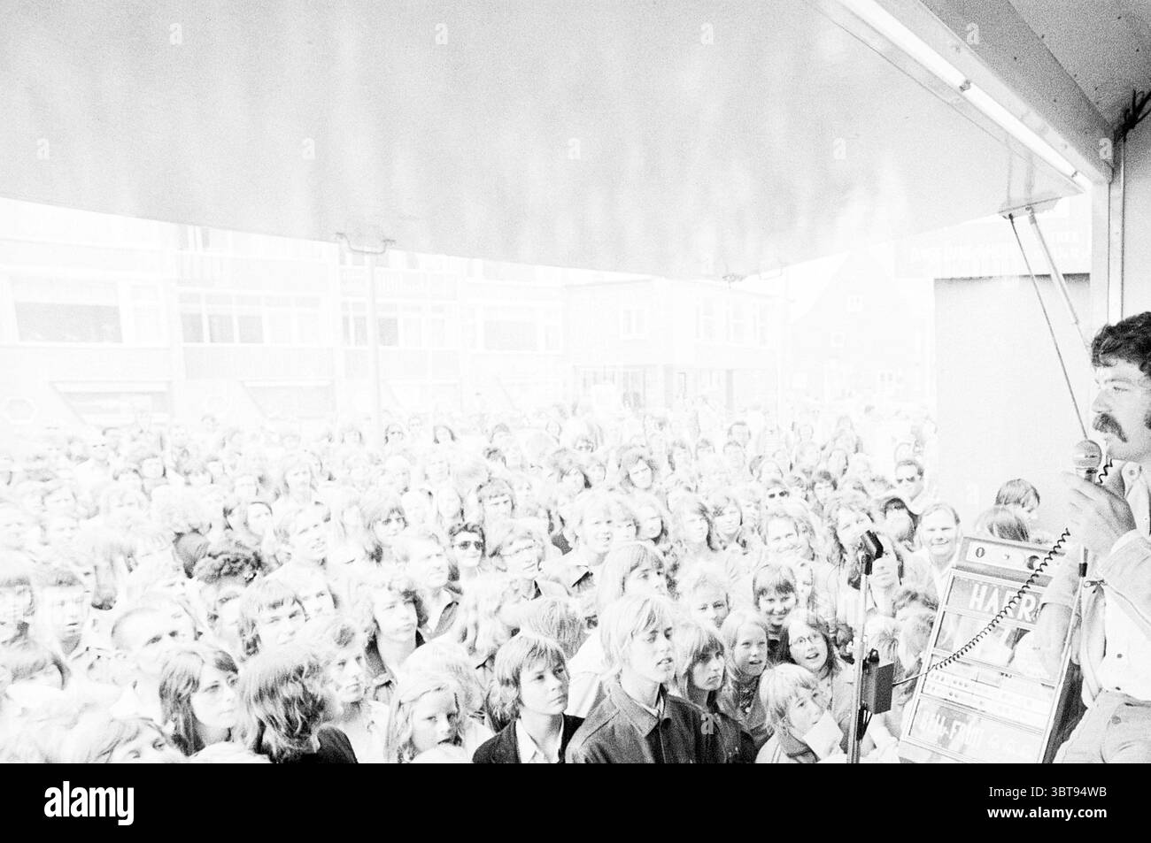 Music Festival, Whizgle News, Dutch Desk, Paesi Bassi, 1950 - 2000 il 15-06-1974. L'immagine contiene questi argomenti. La scena raffigura una grande folla riunita in un ambiente all'aperto, probabilmente per un evento pubblico o una manifestazione. La moltitudine di persone riempie il primo piano, mostrando una varietà di espressioni che vanno dall'eccitazione alla curiosità. Il loro abbigliamento ricorda un'epoca passata, con molti stili caratteristici degli anni '1970, tra cui pantaloni con fondo a campana e camicie a motivi geometrici. Al centro, un altoparlante si trova su una piccola piattaforma, coinvolgendo il pubblico. Appare animato, gestante mentre Foto Stock