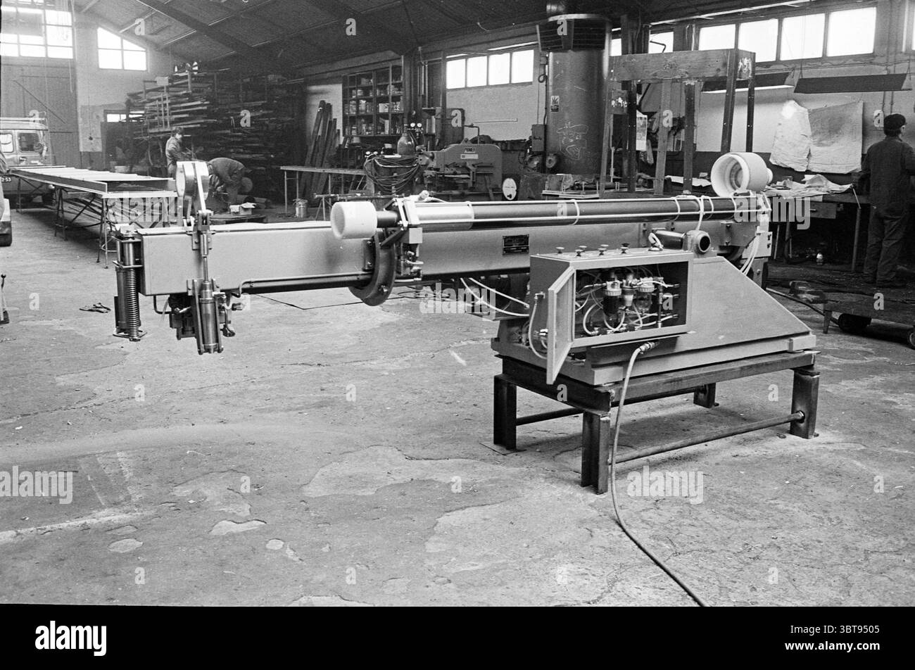 Factory Hall., Whizgle News, Dutch Desk, Paesi Bassi, 1950 - 2000. L'immagine mostra questi argomenti. La scena si svolge in un'ampia bottega caratterizzata da un alto soffitto a volta. La luce naturale si riversa attraverso le grandi finestre, gettando ombre morbide sul pavimento in cemento, che rivela un mix di polvere e usura derivante da anni di utilizzo. In primo piano, una grande macchina industriale si trova in posizione dominante. Il suo robusto telaio è verniciato di grigio tenue, accentuato da componenti metallici usurati che suggeriscono una storia di duro lavoro. La macchina è dotata di varie leve e tubi, suggerendo il suo complesso functio Foto Stock