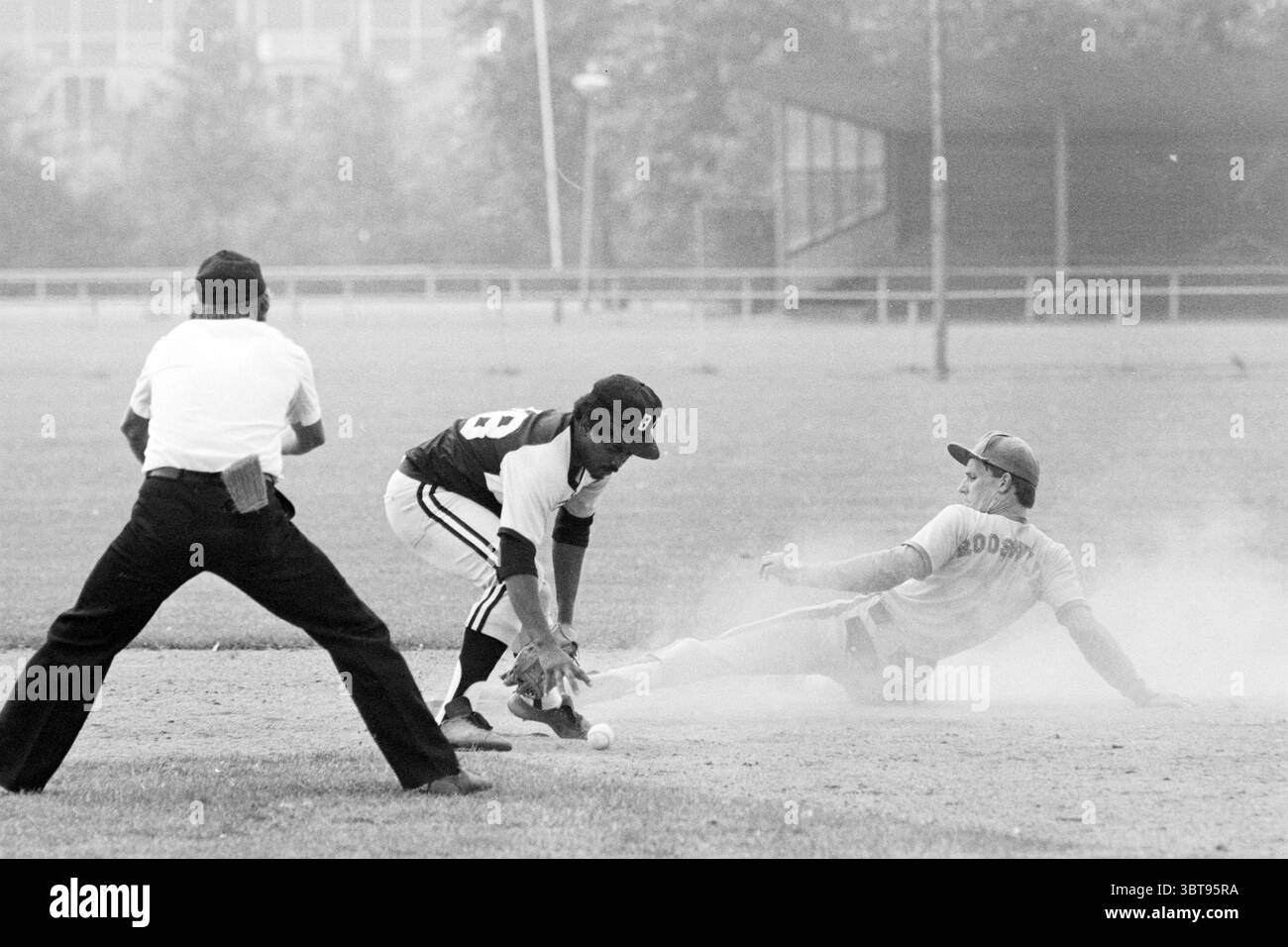 Rooswijk- Black Lions Baseball Rooswijk, Whizgle News, Dutch Desk, Paesi Bassi, 1950 - 2000 il 31-07-1982. Questi argomenti vengono visualizzati nell'immagine. La scena cattura un momento di tensione su un polveroso campo da baseball, dove un gioco si svolge in seconda base. Due giocatori sono al centro dell'attenzione: Uno, vestito con una maglia scura e pantaloni bianchi, accovacciato in posizione difensiva, la sua mano con i guanti che si allunga per toccare una palla da baseball appena alla sua portata. La sua espressione è intensa e focalizzata, sottolineando l'alta posta in gioco. Il giocatore avversario, rivestito in un'uniforme più leggera con toni grigi, sta scivolando verso la seconda base Foto Stock
