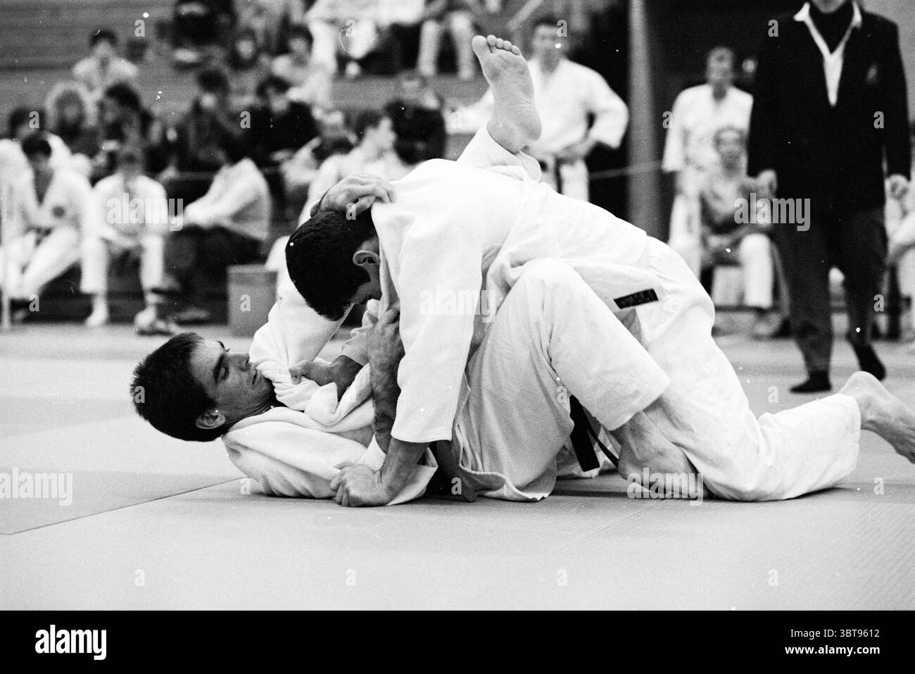 Judo Spaarnehal, Whizgle News, Dutch Desk, Paesi Bassi, 1950 - 2000 il 17-11-1990. Questi sono gli argomenti nell'immagine. La scena cattura un momento intenso durante una partita di judo in una spaziosa palestra piena di spettatori. In primo piano, due concorrenti afferrano un morbido tappeto tatami, il loro judogi bianco che contrasta nettamente con le sfumature del pavimento. Un individuo è a terra, concentrandosi intensamente mentre tenta di sfuggire a una presa, mentre l'altro, posizionato sopra di lui, esercita pressione, mostrando una miscela di forza e tecnica. Intorno ai concorrenti, una folla di persone entusiaste Foto Stock