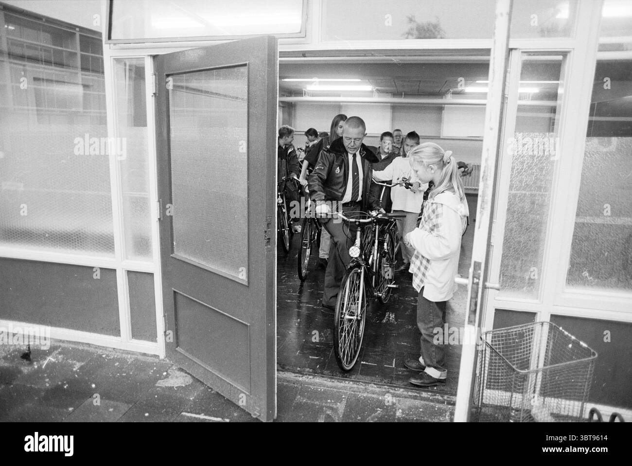 Ispezione bicicletta Ichthus Lyceum, Whizgle News, Dutch Desk, Paesi Bassi, 1950 - 2000 il 25-09-1995. L'immagine contiene questi argomenti. La scena cattura un momento dinamico all'ingresso di una stanza, in cui un gruppo di individui sta muovendo le biciclette. L'ambiente è caratterizzato da un interno spazioso e ben illuminato, con luci fluorescenti che forniscono un'illuminazione luminosa e uniforme che si riflette sul pavimento lucido. In prima linea, un uomo di mezza età, vestito con una giacca scura, si erge accanto a una bicicletta. Si impegna con una ragazza più giovane che lo sta aiutando con impazienza a guidare la moto fuori Foto Stock