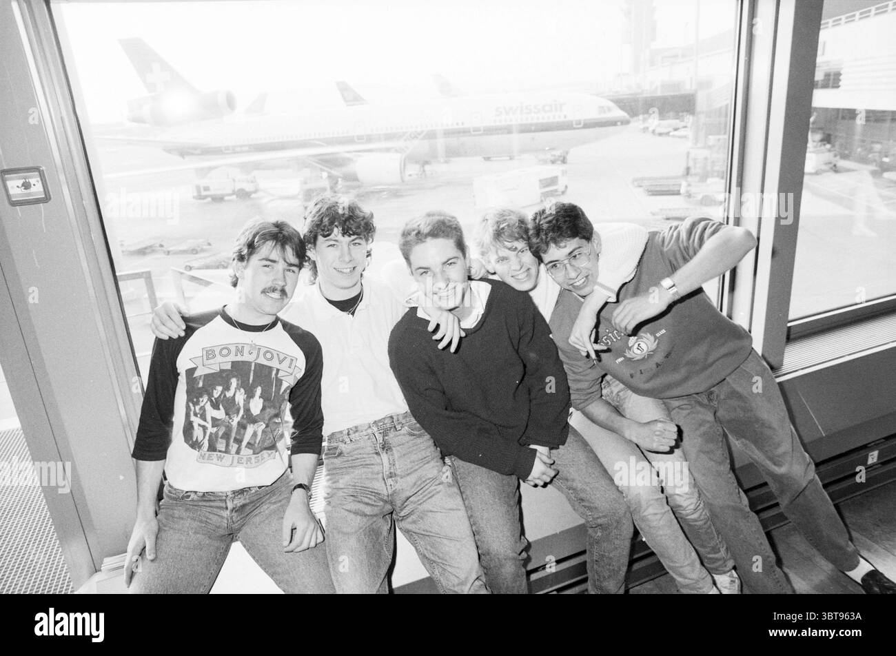 Gita di un giorno a Schiphol - Zurigo Schiphol Flying, Whizgle News, Dutch Desk, Paesi Bassi, tel. 1950 - 2000, tel. 16-12-1988. Questi argomenti vengono visualizzati nell'immagine. Un vivace gruppo di cinque giovani uomini è riunito, seduto su una sporgenza con una grande finestra che offre una vista sulla pista dell'aeroporto. L'ambiente è vivace, con un aereo Swissair visibile all'esterno, che suggerisce viaggi e avventure. Gli individui trasudano un senso di cameratismo, le loro espressioni vanno dai sorrisi giocosi ai granini rilassati. Indossati con un abbigliamento casual, indossano un mix di fantasie e colori: Uno sfoggia una t-shirt grafica con un mu Foto Stock