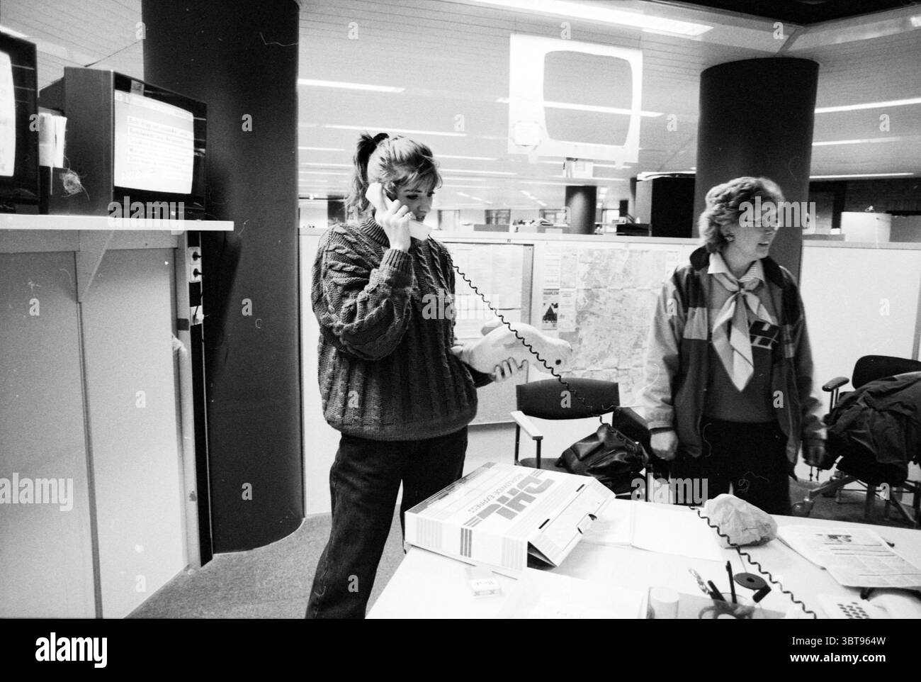 Samantha Minne con animale ripieno, Whizgle News, Dutch Desk, Paesi Bassi, 1950 - 2000 il 17-12-1990. Questi argomenti vengono visualizzati nell'immagine. La scena raffigura un ambiente d'ufficio vivace con particolare attenzione a due donne impegnate in conversazione e lavoro. La donna a sinistra tiene un ricevitore telefonico all'orecchio, la sua espressione suggerisce concentrazione o urgenza mentre comunica. È vestita con un comodo maglione testurizzato, che trasmette un senso di calore in un ambiente altrimenti dai toni neutrali. La donna accanto a lei è leggermente a destra, guardando con un comportamento attento. Lei Foto Stock