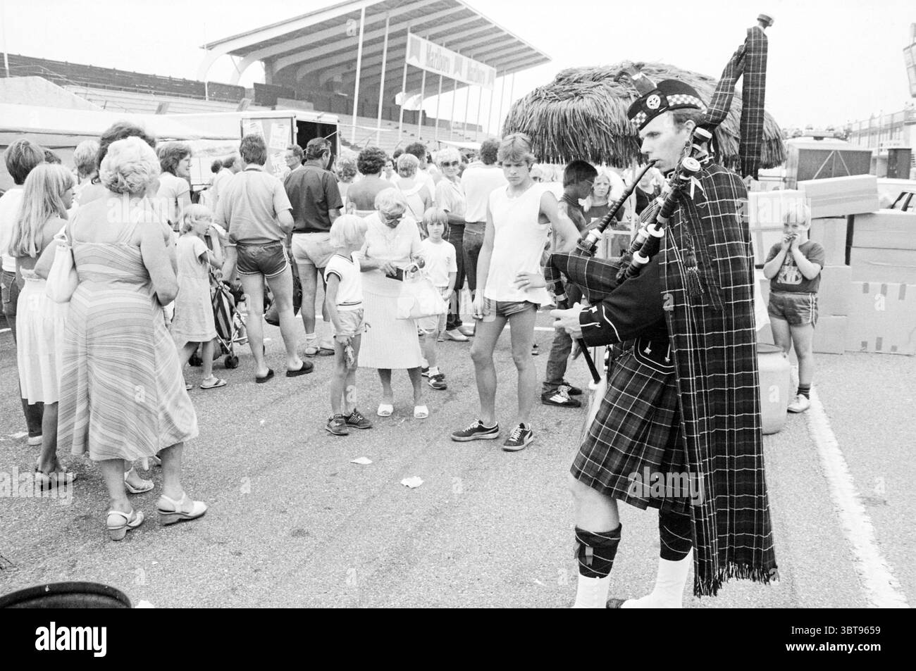 Braderie Circuit Zandvoort Braderie Zandvoort, Whizgle News, Dutch Desk, Paesi Bassi, 1950 - 2000 il 31-07-1982. L'immagine mostra questi argomenti. La scena cattura un vivace evento all'aperto pieno di attività. In primo piano, una persona vestita in tradizionale abbigliamento scozzese, completa di kilt e cappello, suona le cornamuse con intensità espressiva. Il musicista ha una posizione sicura, attirando l'attenzione con il loro elaborato costume, che è adornato con vari elementi decorativi come piume e motivi. Intorno al musicista c'è un gruppo eterogeneo di persone. Alcuni adulti sono enga Foto Stock