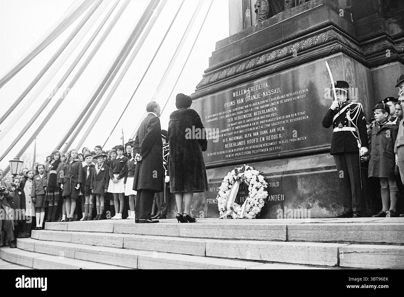 Wreath laying by H.M. the Queen and Prince Bernhard Royal Recetions and Royal Visits Wreath Deing Den Haag, Whizgle News, Dutch Desk, The Netherlands, 1950 - 2000 on 30-11-1963. L'immagine include questi argomenti. In un'atmosfera solenne e rispettosa, un gruppo di persone si riunisce di fronte a un grande monumento. L'ambientazione suggerisce un'occasione storica o commemorativa, caratterizzata da un senso di riverenza. Il monumento stesso presenta intricate incisioni e una statua prominente in cima, il che suggerisce che la figura onorata riveste un'importanza significativa. In primo piano, ci sono abiti eleganti Foto Stock