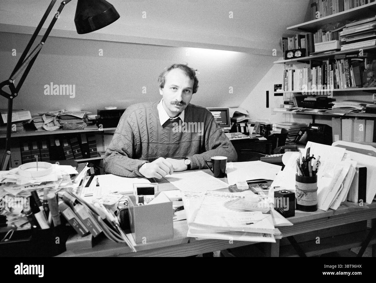 Uomo alla scrivania in una camera attico con una pila di riviste Personal computer Magazine, Whizgle News, Dutch Desk, Paesi Bassi, 1950 - 2000 su 12-1989. L'immagine contiene questi argomenti. La scena presenta un'area di lavoro ingombrante piena di documenti, libri e varie forniture per ufficio. Un uomo siede su una scrivania di legno, la sua espressione è premurosa mentre guarda direttamente lo spettatore. L'uomo ha i capelli castani chiari, i baffi e indossa un comodo maglione in maglia a cavo dai toni tenui. Intorno alla scrivania c'è una disposizione caotica di documenti, quaderni e cartelle, che suggerisce un ambiente vivace e creativo. Un bla Foto Stock