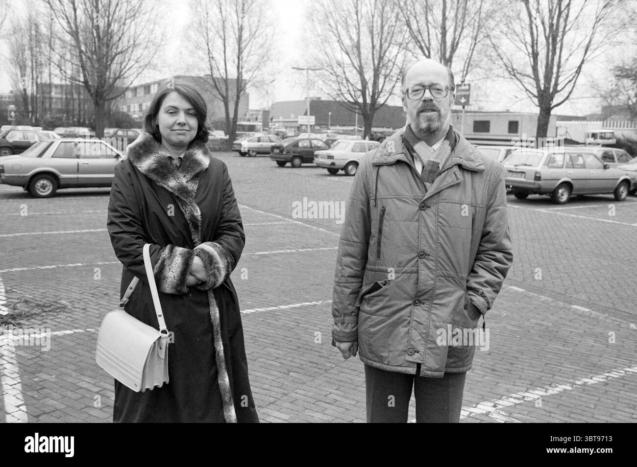 Aldermen Cnossen e Vrij-Vringer Haarlemmermeer in un parcheggio Hoofddorp Paesi Bassi, Whizgle News, Dutch Desk, Paesi Bassi, 1950 - 2000 il 22-11-1985. Questi sono gli argomenti nell'immagine. La scena è ambientata in un ampio parcheggio aperto, pieno di una varietà di veicoli che suggeriscono una miscela di modelli più vecchi e design più moderni. Il terreno è composto da mattoni pavimentati scuri, che sottolineano la sofferenza dell'ambiente. In primo piano, due persone stanno vicini. La prima persona è una donna dall'espressione seria, vestita con un lungo cappotto scuro adornato da una colla di pelliccia Foto Stock