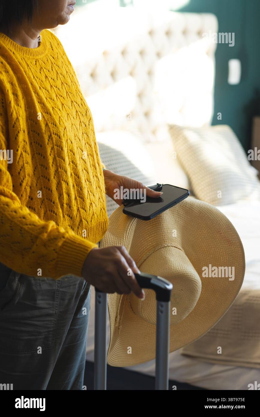 Donna che afferra la valigia a letto in camera tenendo in mano il cappello di paglia e lo smartphone, spazio per copiare Foto Stock