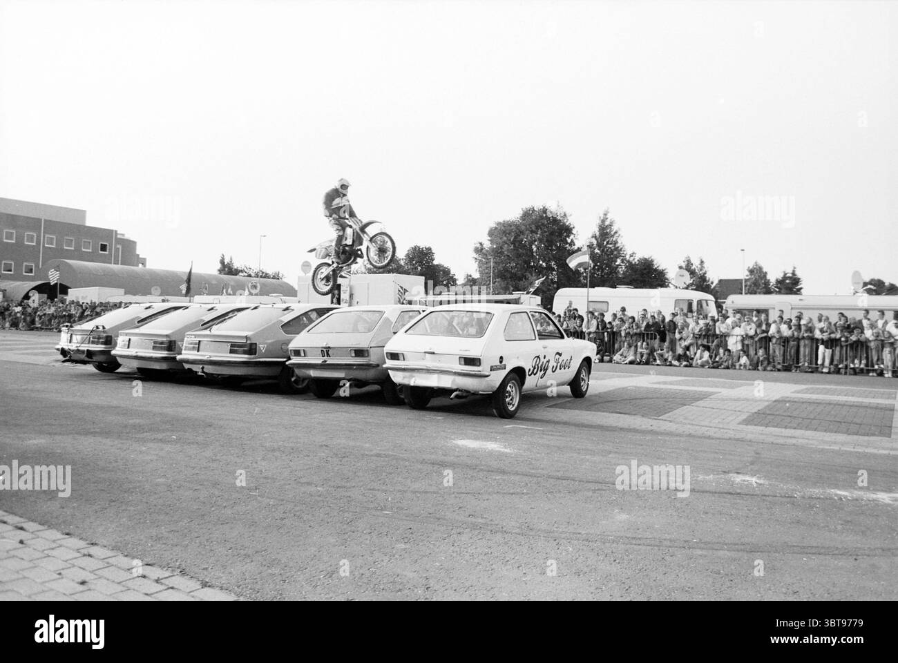 Spettacolo acrobatico Big Foot Cars Hoofddorp Paesi Bassi, Whizgle News, Dutch Desk, Paesi Bassi, 1950 - 2000 il 19-06-1992. L'immagine contiene questi argomenti. La scena cattura un momento emozionante in un evento acrobatico all'aperto. In primo piano, diverse auto compatte sono allineate in fila, parcheggiate vicino con i loro cofani rivolti verso l'esterno. I veicoli sono verniciati in modo uniforme in tonalità di luce e scura, contribuendo a creare un tema monocromatico, sottolineando ulteriormente l'azione dinamica che si verifica sopra di essi. Al centro dell'immagine, un pilota di motocross con un casco e un equipaggiamento protettivo completo salta dentro Foto Stock