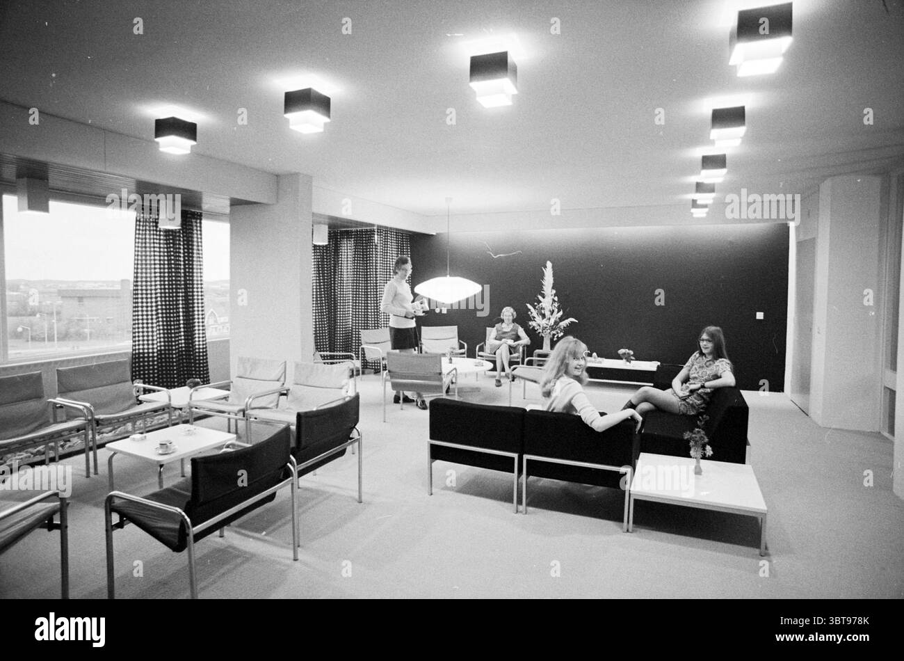 INTR. Huis ter Wijck Interior, Whizgle News, Dutch Desk, Paesi Bassi, 1950 - 2000 il 18-11-1971. L'immagine contiene questi argomenti. La scena raffigura un interno moderno e minimalista, caratterizzato da una spaziosa area lounge con un ambiente sofisticato e un po' retrò. La sala è illuminata da una serie di plafoniere rettangolari che emettono una luce soffusa e diffusa, creando un'atmosfera calda ma discreta. Nel centro, diversi gruppi di persone sono organizzati casualmente. Un uomo sta vicino a una finestra, impegnato in una conversazione, mentre due donne sono sedute su un elegante divano nero. Una donna si appella Foto Stock