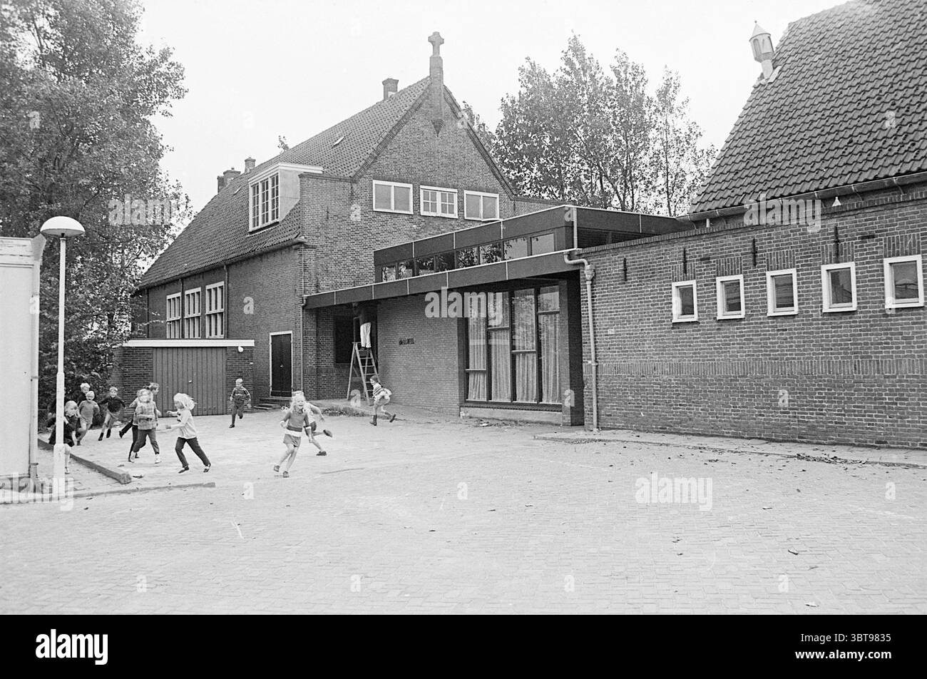 Esterno della scuola materna Luifel, Whizgle News, Dutch Desk, Paesi Bassi, 1950 - 2000. Questi sono gli elementi dell'immagine. La scena raffigura un edificio educativo della metà del XX secolo caratterizzato dalla sua architettura distinta. La struttura presenta una combinazione di mattoni e elementi moderni, tra cui grandi finestre che consentono alla luce naturale di entrare. La facciata presenta un mix di tetti inclinati tradizionali e superfici piane contemporanee, sottolineando sia la storia che il progresso. In primo piano, un gruppo di bambini si impegna in attività ludiche, sparse per il cortile. Loro Foto Stock