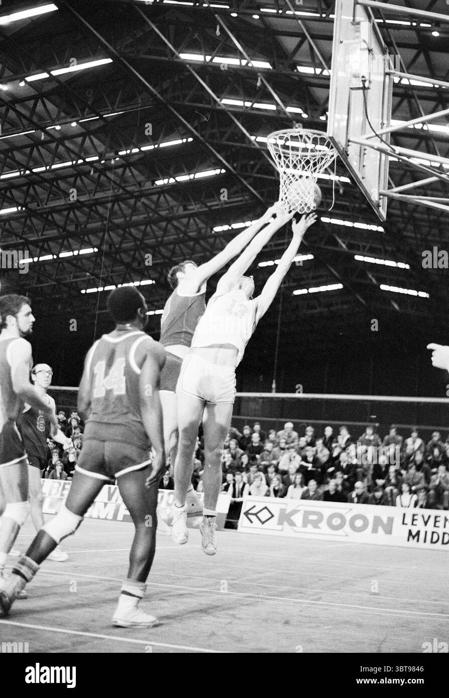 Partita di basket maschile, Whizgle News, Dutch Desk, Paesi Bassi, 1950 - 2000 il 12-1969. L'immagine mostra questi argomenti. La scena cattura un momento intenso in una partita di basket, ambientata all'interno di una grande arena al coperto. L'alto soffitto è sostenuto da una complessa griglia di travi metalliche che creano ombre geometriche sul pavimento in legno duro. Il campo è illuminato da luci luminose, creando un forte contrasto tra aree chiare e scure. In primo piano, due giocatori sono impegnati in una lotta dinamica vicino al canestro da basket. Un atleta, che indossa un'uniforme bianca con il numero 13, sta saltando in aria Foto Stock