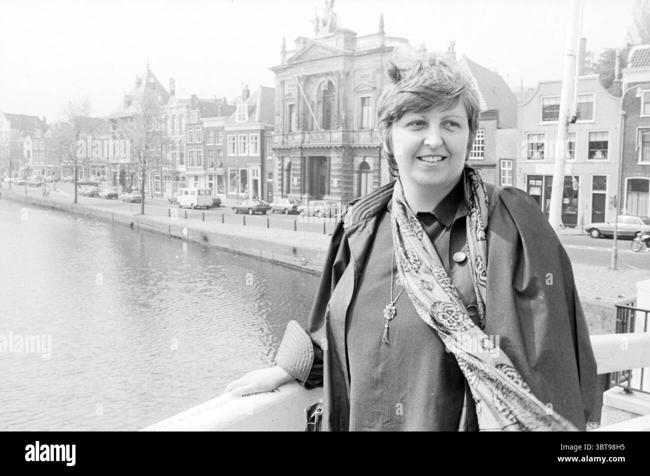 Sig.ra Romé Defour per Teylers Museum Museum Museum Museum Museums People Haarlem Nederland, Whizgle News, Dutch Desk, Paesi Bassi, 1950 - 2000 il 04-05-1984. L'immagine mostra questi argomenti. In un ambiente urbano sereno, una donna si erge con fiducia sul lungofiume, affacciata su un calmo corpo d'acqua. L'architettura che la circonda trasuda fascino storico, con eleganti edifici che costeggiano la sponda opposta, sfoggiando una varietà di stili dal classico al moderno. La donna è vestita con un outfit dalle tonalità scure completato da una sciarpa a fantasia drappeggiata elegantemente intorno al collo. I suoi capelli corti sono curati in modo informale, aggiungendo t Foto Stock