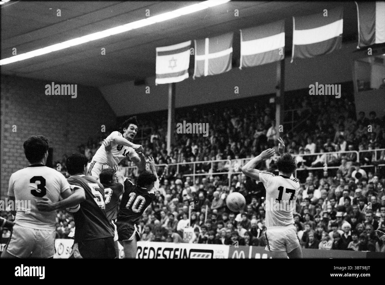 Paesi Bassi - Israel Handball, Whizgle News, Dutch Desk, Paesi Bassi, 1950 - 2000 il 02-03-1983. L'immagine mostra questi argomenti. La scena cattura un momento intenso in un'arena sportiva, probabilmente durante una partita di pallamano. L'atmosfera è carica di energia mentre gli atleti sono impegnati in movimenti dinamici. In primo piano, i giocatori sono in varie posizioni, mostrando il loro atletismo. Un giocatore vestito con una maglia bianca (numero 1) salta in aria, mostrando un potente salto mentre si prepara a lanciare la palla. La sua espressione è concentrata e determinata, trasmettendo l'intensità della competizione. Foto Stock
