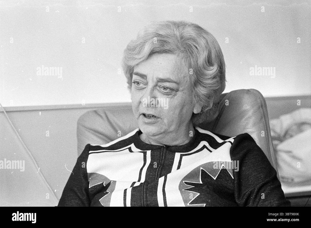 Mrs. Van der Ham Haarlem Persons Portraits, Whizgle News, Dutch Desk, Paesi Bassi, 1950 - 2000 il 07-12-1973. Questi argomenti sono mostrati nell'immagine. In questa scena, una donna siede su una sedia confortevole e strutturata con una postura rilassata. Ha capelli ondulati e di media lunghezza che sono stati delicatamente modellati, creando un contorno elegante intorno al suo viso. L'illuminazione è morbida e diffusa, fornendo una luce soffusa che mette in risalto le sue caratteristiche facciali senza ombre. Il suo abbigliamento consiste in un capo scuro impreziosito da un vivace motivo geometrico al centro, caratterizzato da linee nette e colori contrastanti Foto Stock