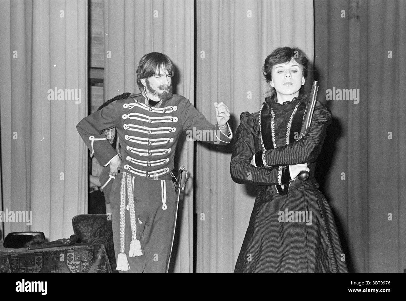 Gli studenti di Klaas de Vriesschool suonano Chekhov Schools Theater, Whizgle News, Dutch Desk, Paesi Bassi, 1950 - 2000 il 08-02-1977. Questi argomenti sono mostrati nell'immagine. In un ambiente teatrale, due individui sono il punto focale della scena. A sinistra, un uomo vestito con un'uniforme militare adornata da strisce distinte sembra essere in una posa animata, probabilmente a metà azione. Il suo outfit è prevalentemente scuro con tocchi bianchi che creano un contrasto audace. L'espressione sul suo volto trasmette entusiasmo o intensità, riflettendo la natura drammatica della performance. Da a t Foto Stock