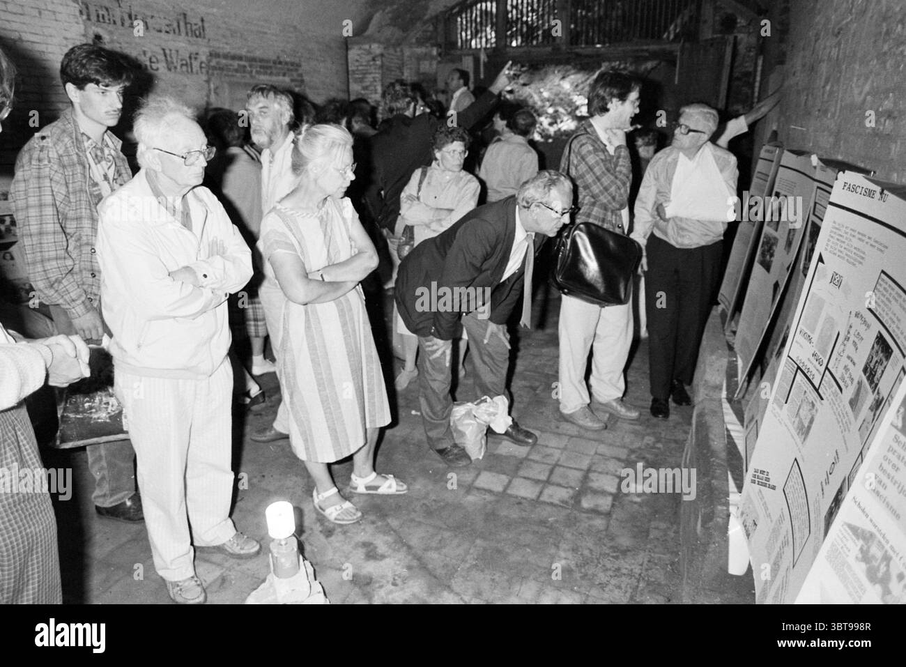 Fort Island IJmuiden IJmuiden Velsen IJmuiden Paesi Bassi, Whizgle News, Dutch Desk, Paesi Bassi, 1950 - 2000 il 20-05-1989. L'immagine mostra questi argomenti. La scena si svolge in uno spazio sotterraneo scarsamente illuminato, possibilmente una cantina rustica o una sala espositiva con pareti in mattoni a vista. Un gruppo di persone è riunito, le loro espressioni riflettono la curiosità e l'impegno. La folla è costituita principalmente da uomini e donne di varie età, ognuno con stili distinti nei propri abiti. Gli anziani, uno con occhiali e capelli grigi, stanno vicini, mentre un uomo di mezza età si appoggia Foto Stock