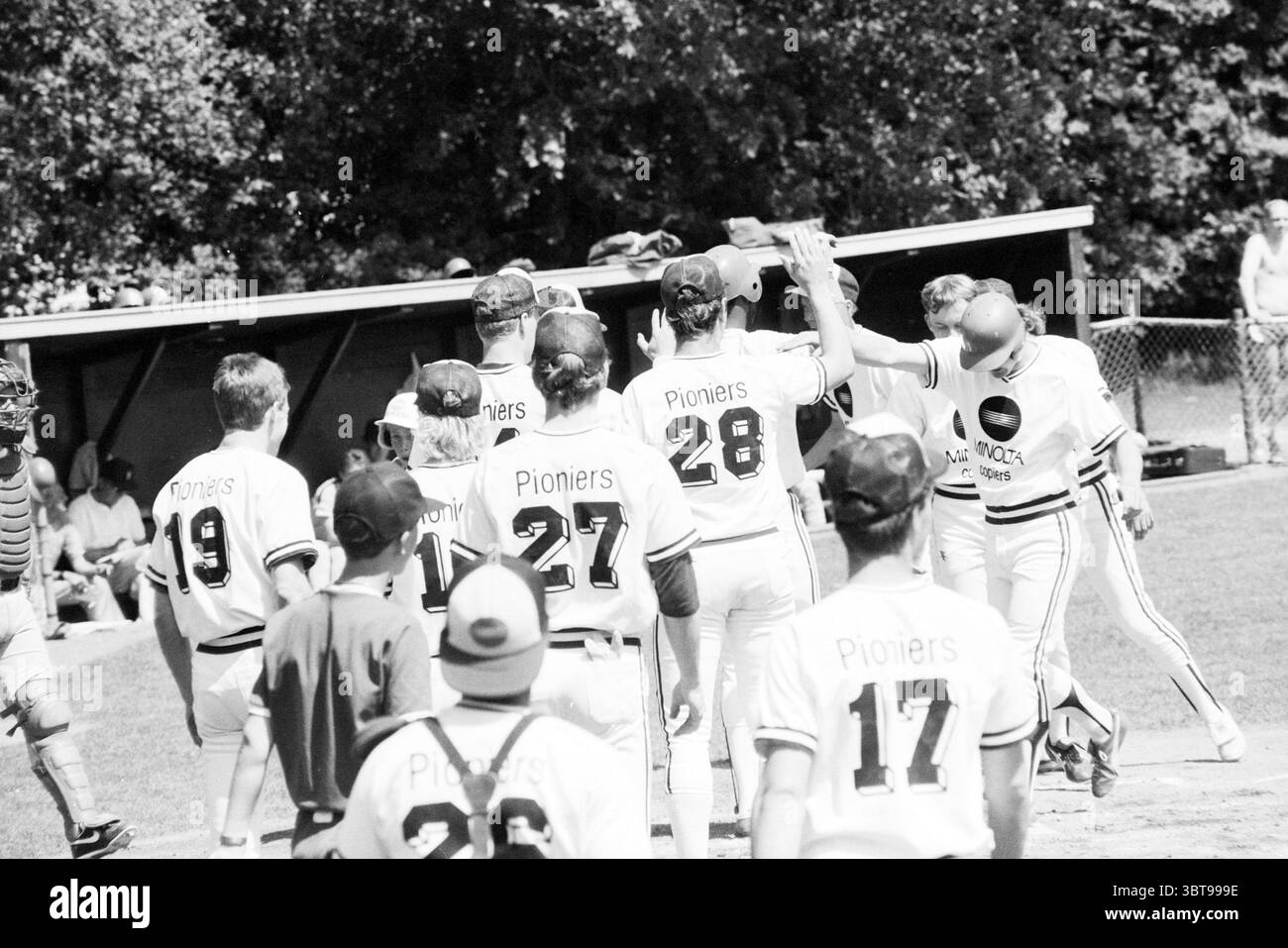 Baseball Pioneers - Amstel Tigers, Whizgle News, Dutch Desk, Paesi Bassi, 1950 - 2000 nel 1986. L'immagine include questi argomenti. La scena cattura un momento su un campo da baseball, dove un gruppo di giocatori con uniformi abbinate, prevalentemente bianche con distintivi accenti neri e rossi, festeggiano. Queste uniformi portano il nome "pionieri", che indica uno spirito di squadra e una cameratismo tra i giocatori. In primo piano, sono riuniti diversi giocatori, alcuni con cappelli rivolti all'indietro, suggerendo un'atmosfera rilassata e informale. Alcuni giocatori sono già in procinto di scontrarsi a vicenda, Th Foto Stock