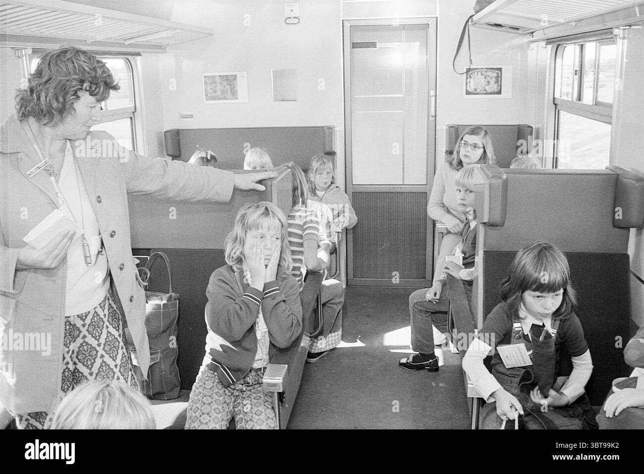 Bambini in treno per Rotterdam, Whizgle News, Dutch Desk, Paesi Bassi, 1950 - 2000 nel 1973. Questi argomenti sono mostrati nell'immagine. In una carrozza ferroviaria d'epoca, un gruppo di bambini è seduto tra gli elementi interni familiari di un pullman ferroviario. La tavolozza dei colori è silenziata, con sfumature di grigio e beige che dominano la scena. I posti a sedere sono disposti a coppie, con morbide panchine imbottite che creano uno spazio intimo, anche se leggermente ridotto. Da un lato, una donna adulta, forse insegnante o chaperone, si erge mentre gestiva un bambino davanti. Ha i capelli ricci e indossa un colore chiaro Foto Stock
