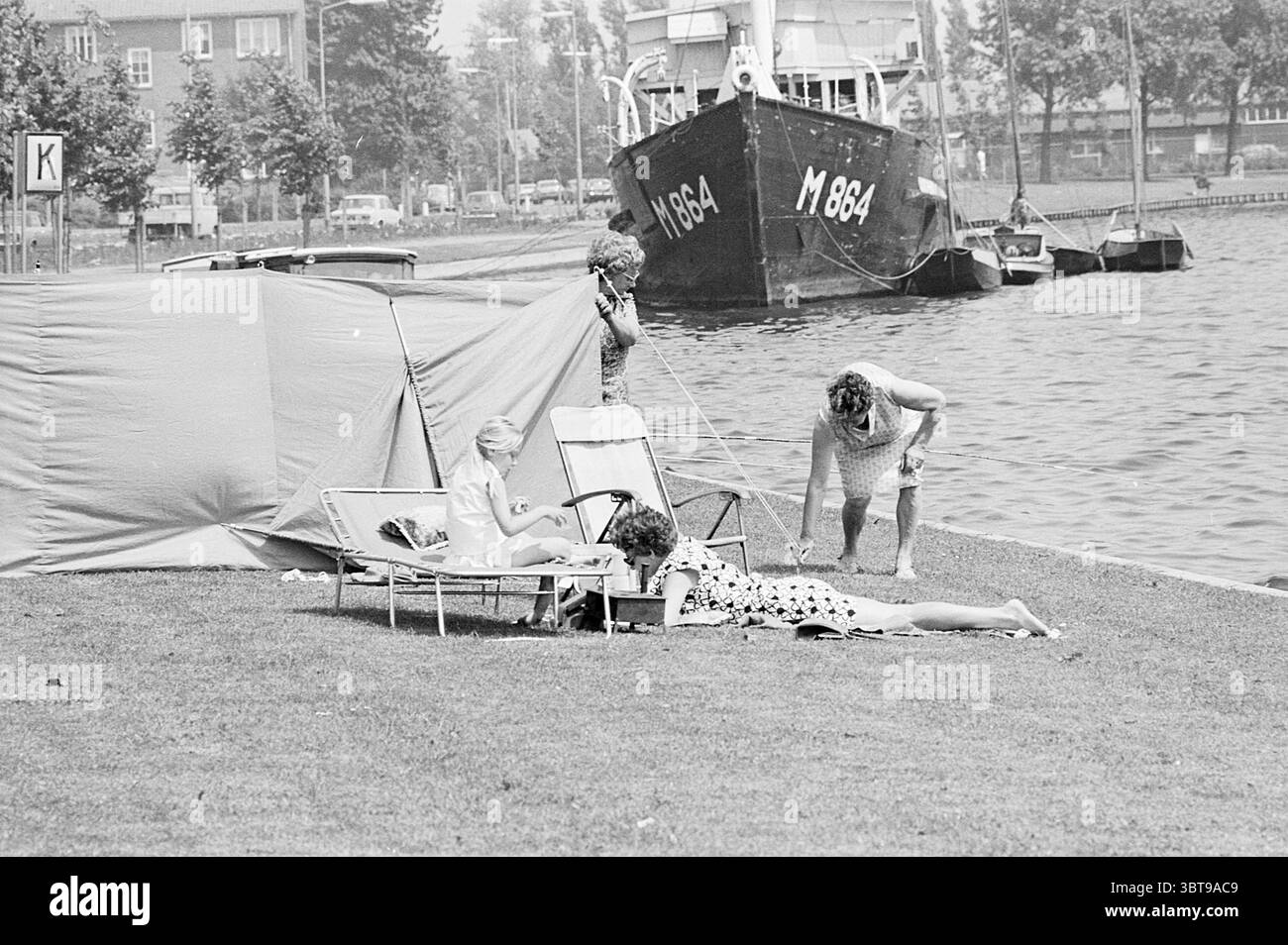 Solarium per signore, Whizgle News, Dutch Desk, Paesi Bassi, 1950 - 2000. L'immagine include questi argomenti. La scena raffigura un tranquillo ambiente lungo il fiume, caratterizzato da un fiorente prato verde che si trasforma in acqua sul bordo. Due persone si stanno rilassando in primo piano: Una è sdraiata sull'erba, vestita con un abito a motivi di colore chiaro, mentre l'altra è seduta su una poltrona, impegnata in qualche attività. La loro postura informale suggerisce un pomeriggio rilassato. Nelle vicinanze si trova una grande tenda sul prato, che fornisce ombra e il suo tessuto cattura la leggera brezza. Il surf della tenda Foto Stock