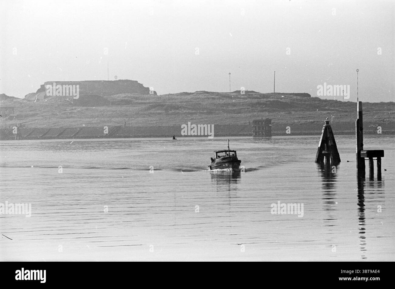 Foto del porto di IJmuiden Ships, Whizgle News, Dutch Desk, Paesi Bassi, 1950 - 2000 il 17-02-1961. L'immagine include questi argomenti. In questa scena, un sereno corso d'acqua si estende attraverso il primo piano, la sua superficie riflette i gradienti di luce morbidi. L'acqua sembra calma e ferma, creando un effetto simile a un vetro che rispecchia l'ambiente circostante. Una piccola barca naviga attraverso l'acqua, seguendo un dolce risveglio dietro di essa, suggerendo movimento e scopo. All'orizzonte si innalza un dolce pendio, adornato di erba e possibilmente arbusti bassi, che lasciano intendere un paesaggio naturale. Una struttura simile a un forte è P. Foto Stock