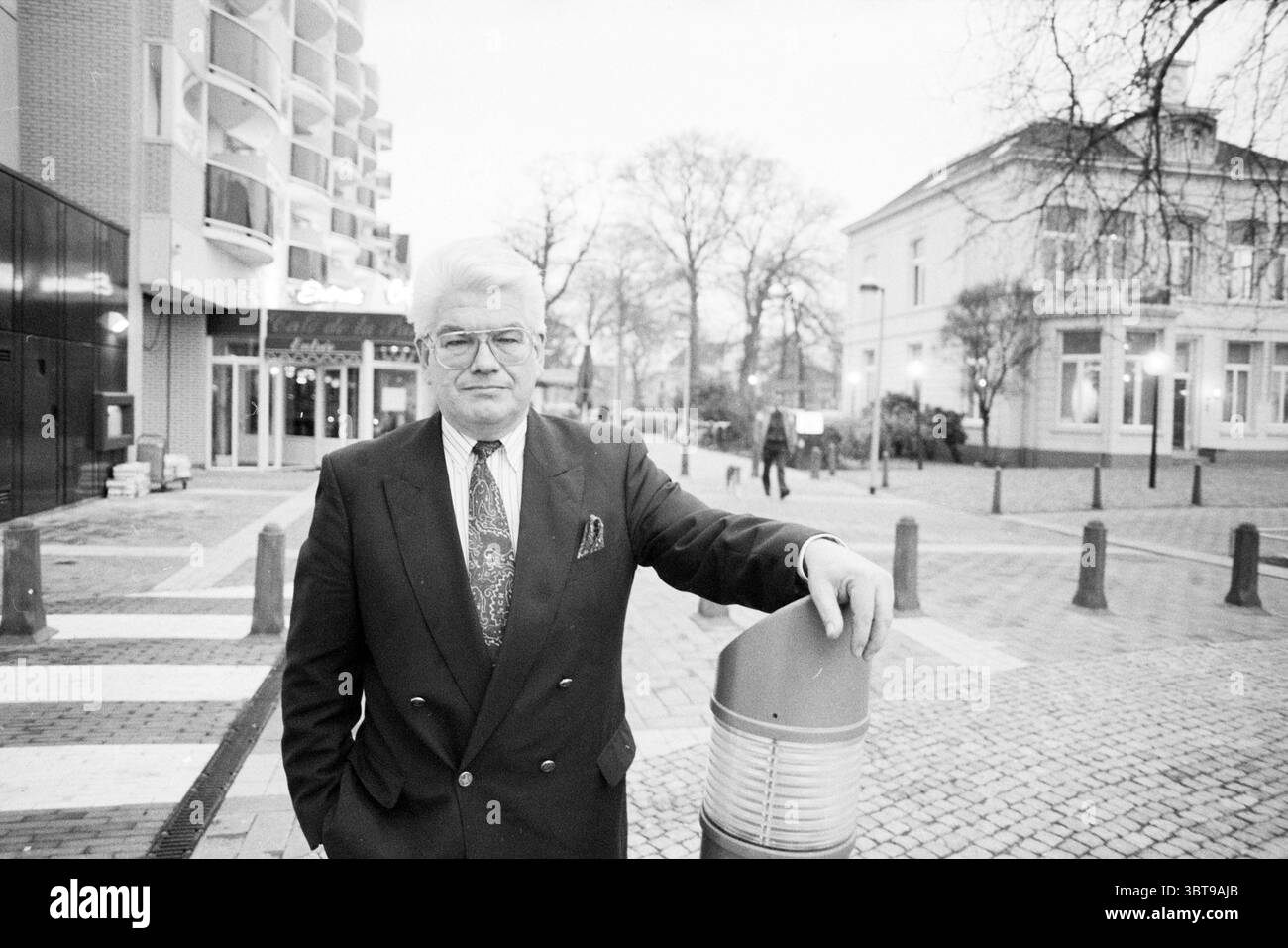 Sig. Davids nuovo presidente KNOV Associations, Whizgle News, Dutch Desk, Paesi Bassi, 1950 - 2000 il 10-01-1990. L'immagine contiene questi argomenti. In un vivace ambiente urbano, un uomo di mezza età si erge con sicurezza su un passaggio pedonale pavimentato. È vestito con un abito scuro, i revers ben definiti, con una cravatta a motivi geometrici che aggiunge un tocco di raffinatezza. L'uomo ha i capelli ben disegnati, incorniciati da un paio di occhiali che enfatizzano il suo comportamento. Intorno a lui ci sono elementi di una vivace scena di strada. A sinistra, un edificio moderno con una facciata in vetro contrasta con il più antico, più tradizionale archite Foto Stock
