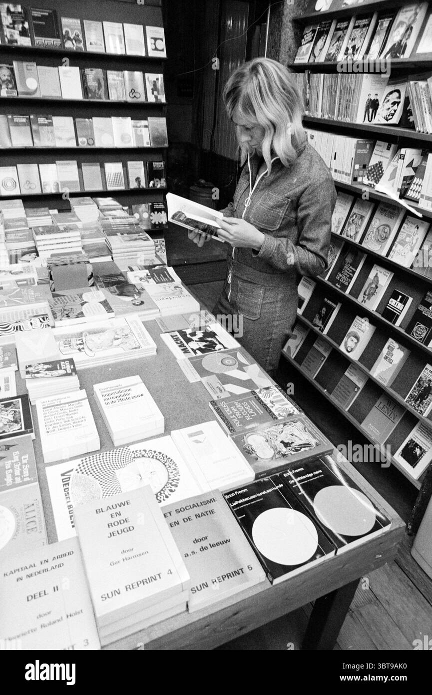 Report Eating Fries and Books Report, Whizgle News, Dutch Desk, Paesi Bassi, 1950 - 2000 il 19-08-1972. Questi argomenti vengono visualizzati nell'immagine. La scena è ambientata in un'accogliente libreria, caratterizzata da numerosi scaffali pieni di una collezione diversificata di libri in vari colori e dimensioni. Gli scaffali sono ben chiusi, mostrando una vasta gamma di titoli che suggeriscono un'attenzione sia alla letteratura che all'arte, data l'eclettico mix di copertine. In primo piano, una donna si trova su un grande tavolo di legno pieno di libri. È vestita con un elegante outfit grigio che aggiunge un tocco di raffinatezza Foto Stock