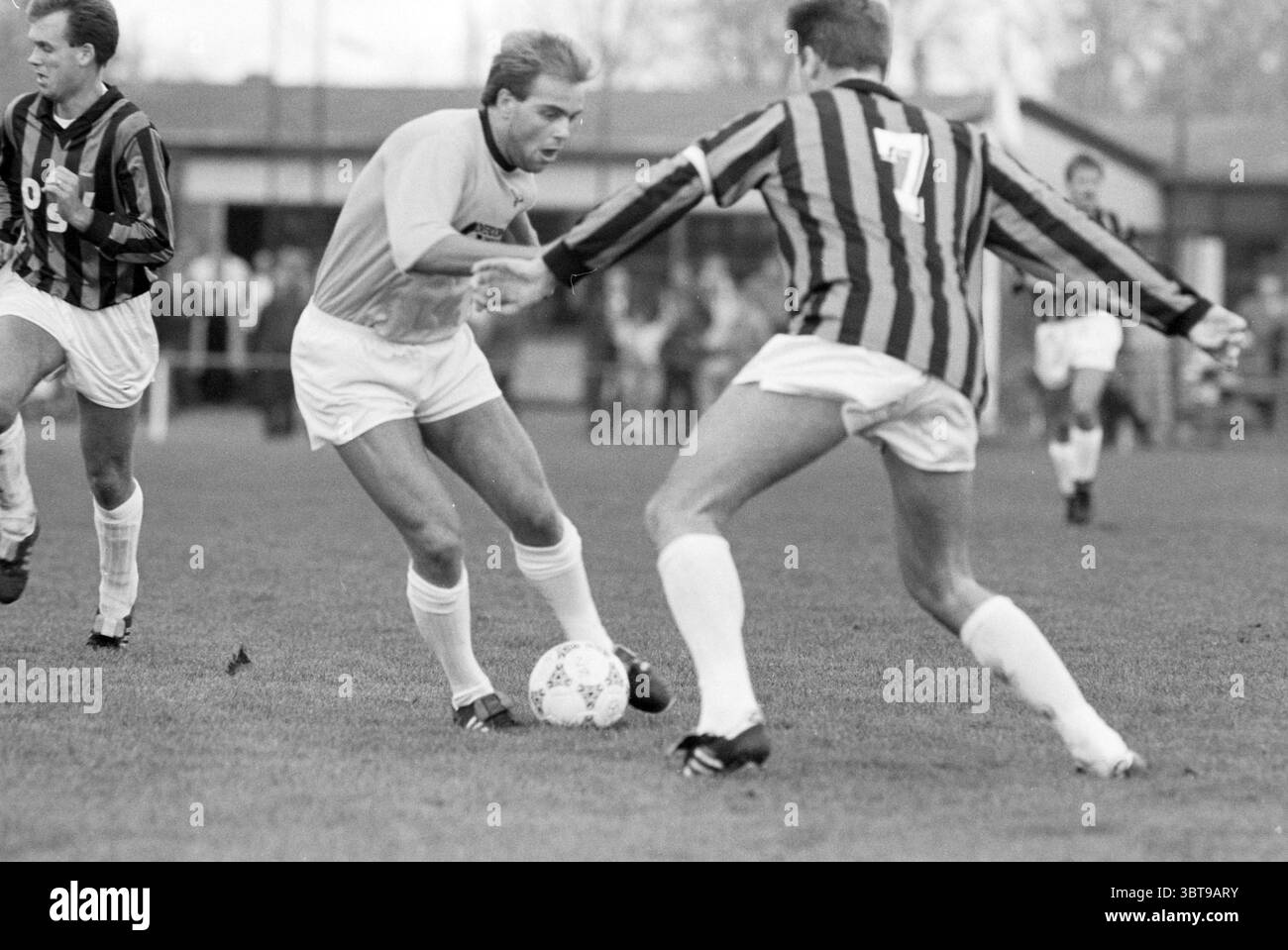 Football Fulmars - OSV, Whizgle News, Dutch Desk, Paesi Bassi, 1950 - 2000 il 03-11-1990. Questi argomenti sono mostrati nell'immagine. La scena raffigura un momento dinamico su un campo da calcio, in cui due giocatori sono intensamente impegnati in un gioco competitivo. Un giocatore, vestito con un'uniforme di colore chiaro, sta sfrecciando la palla sapientemente, mostrando concentrazione e agilità. I suoi capelli sono modellati con cura e sul viso è incisa una leggera determinazione mentre muove la palla intorno all'avversario. Il giocatore avversario, rivestito con una divisa scura a righe con un numero audace impresso sulla schiena, è in un defensi Foto Stock