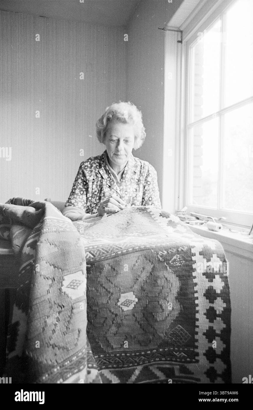 Sig.ra [...] riparatore di tappeti Zandvoort Persons, Whizgle News, Dutch Desk, Paesi Bassi, 1950 - 2000 il 09-10-1964. Questi argomenti vengono visualizzati nell'immagine. Una donna siede ad un tavolo di legno, coinvolta nel suo lavoro. Ha capelli chiari e ondulati con cura e il suo abbigliamento presenta una blusa a motivi floreali che contrasta delicatamente con le tonalità circostanti. Il punto focale della sua attività è una coperta riccamente intrecciata a mano drappeggiata sul tavolo, che mostra intricati disegni geometrici in colori terrosi: Marrone, rosso e bianco. I modelli suggeriscono un legame con la tradizione e gli artigiani Foto Stock