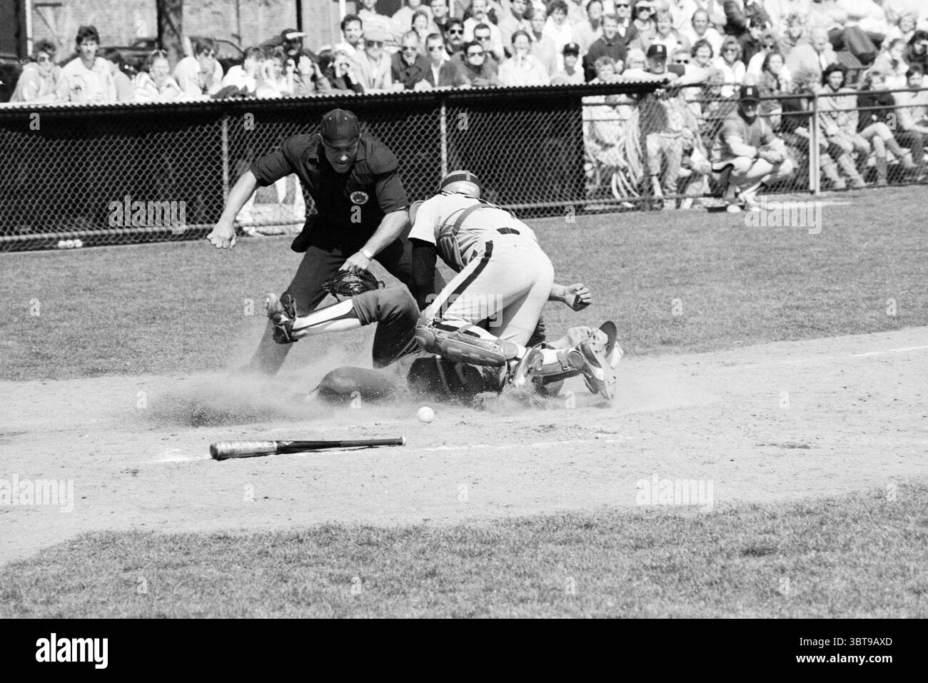 Kinheim - Nicols Baseball Kinheim, Whizgle News, Dutch Desk, Paesi Bassi, 1950 - 2000 il 24-05-1986. Questi sono gli argomenti nell'immagine. La scena cattura un momento intenso durante una partita di baseball, segnato da una collisione dinamica sul piatto di casa. In primo piano, un ricevitore, completamente equipaggiato con equipaggiamento protettivo, è accovacciato a terra, colori regionali della sua uniforme in contrasto con la polverosa e marrone interno. La sua espressione, sebbene oscurata dalla maschera del casco, suggerisce una miscela di determinazione e concentrazione. Accanto a lui, un corridore di base è a metà azione, si tuffa verso casa, gli arti si allungano Foto Stock