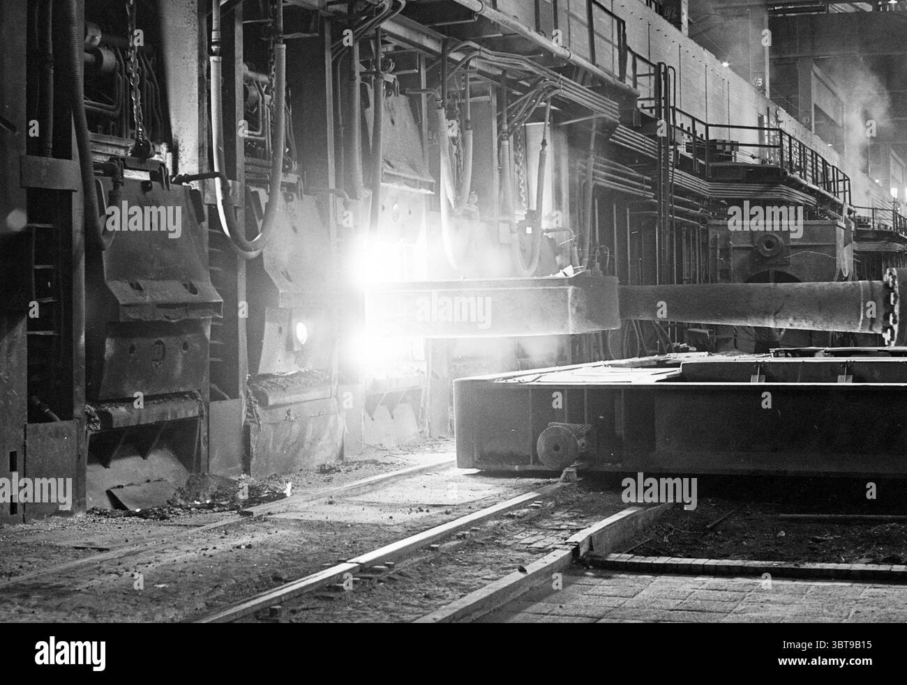 Sciogliendo gli stiletti arresi armi stemmi familiari Velsen-Noord Hoogovens, Whizgle News, Dutch Desk, Paesi Bassi, 1950 - 2000 il 30-08-1965. Questi argomenti vengono visualizzati nell'immagine. La scena è ambientata in un ambiente industriale, caratterizzato da macchinari pesanti e da un senso di attività che suggerisce un impianto di produzione o di trasformazione. I colori dominanti sono sfumature di grigio e nero, che conferiscono un'atmosfera acuta e grintosa che riflette le condizioni di lavoro tipiche di un luogo del genere. In primo piano, una grande struttura metallica pesante è prominente, probabilmente un pezzo di macchinari o attrezzature utilizzati in Foto Stock