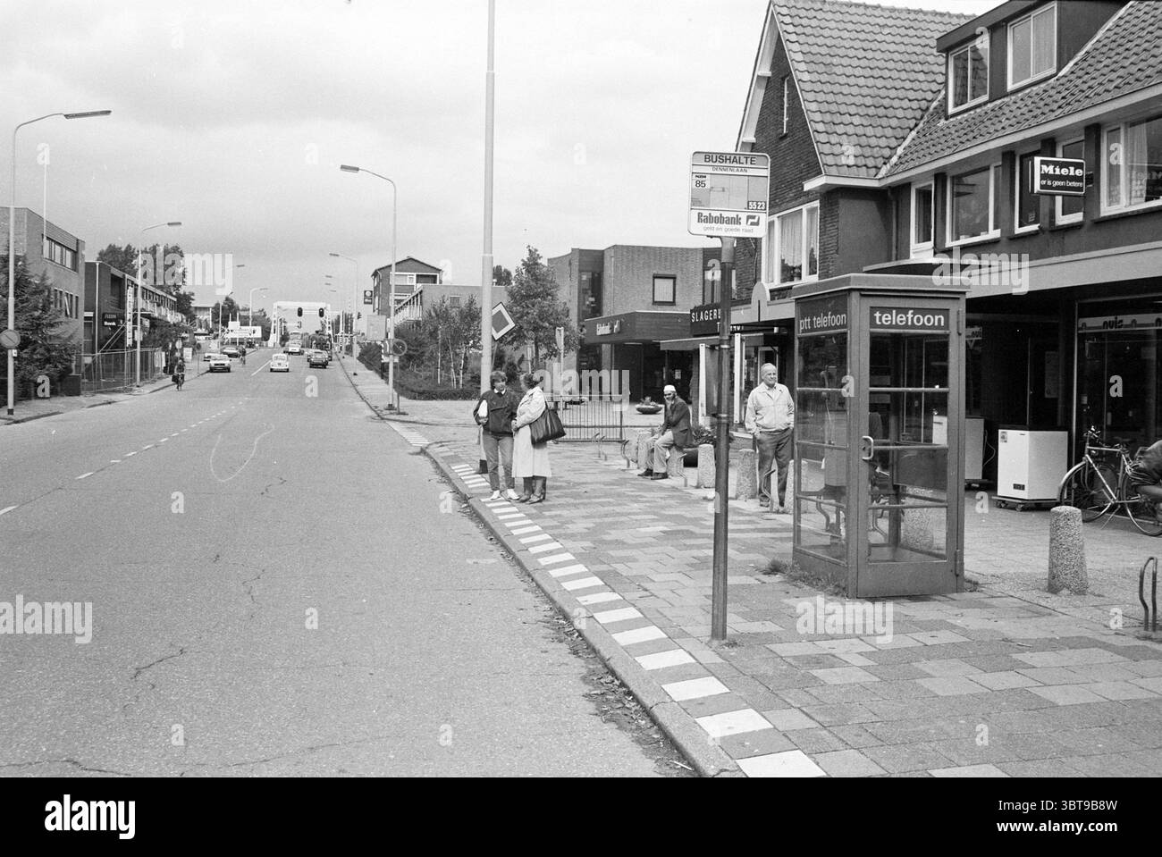 Tour Halfweg ingresso CSM con cigno Halfweg Halfweg, Whizgle News, Banco olandese, Paesi Bassi, 1950 - 2000 il 04-09-1985. L'immagine mostra questi argomenti. La scena cattura un ambiente di strada che trasmette un senso di calma vita suburbana. Un'ampia strada asfaltata attraversa il centro, conducendo l'occhio verso un incrocio distante. Da un lato, diverse persone aspettano tranquillamente presso una fermata dell'autobus designata, le loro posture accennano a un mix di impazienza e conversazione informale. Gli individui sono vestiti con abiti di tutti i giorni, riflettendo vari stili ed età, suggerendo una comunità diversificata. Stanno su un patte Foto Stock