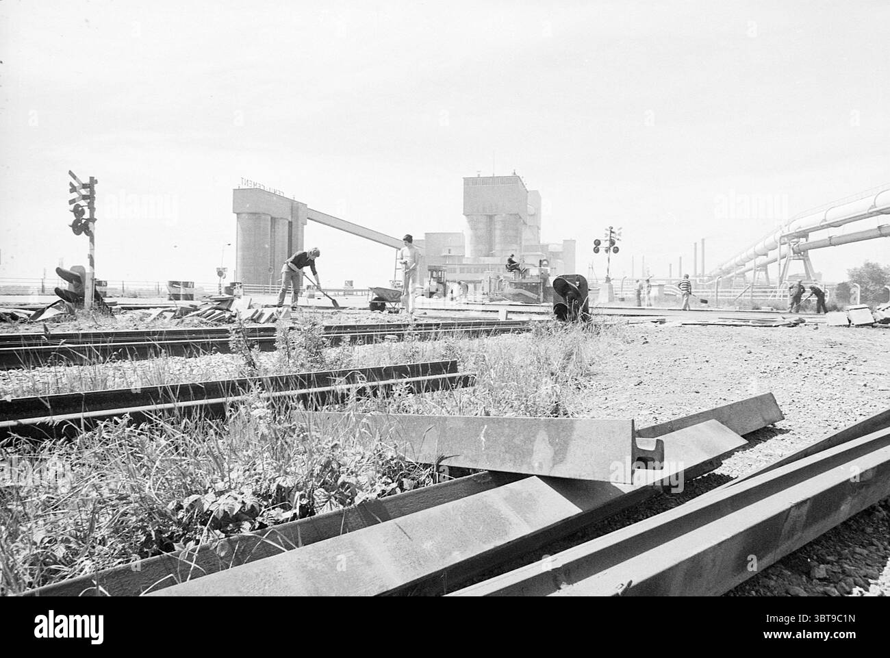 Attraversamento ferroviario a Cemij IJmuiden Railways, Whizgle News, Dutch Desk, Paesi Bassi, 1950 - 2000 su 26-06-1977. Questi argomenti sono mostrati nell'immagine. La scena raffigura un ambiente di lavoro dominato da elementi industriali. In primo piano, diverse lunghezze di binari ferroviari in acciaio si estendono attraverso il terreno, parzialmente ricoperti di erbacce e erbe, sottolineando una miscela di natura e industria. I binari si estendono in direzioni divergenti, accentuando la complessità del sito. I lavoratori sono impegnati in varie attività lungo i binari. Un uomo, inginocchiato, sembra stia ispezionando Foto Stock
