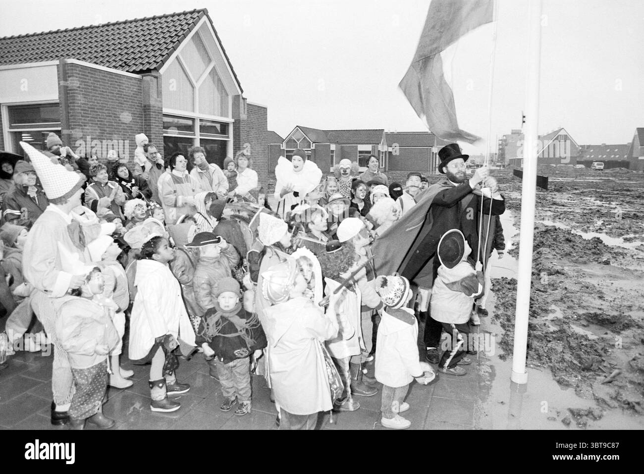 Apertura della nuova scuola De Regenboog a Overbos Hoofddorp apertura inaugurazione scuole Hoofddorp Nederland, Whizgle News, Dutch Desk, Paesi Bassi, 1950 - 2000 il 31-01-1988. Questi argomenti vengono visualizzati nell'immagine. In una vivace scena all'aperto, un grande gruppo di bambini e adulti si riuniscono intorno a un flagpole, alcuni dei quali indossano costumi festivi e abiti colorati. I bambini mostrano una varietà di emozioni espressive, che vanno dall'eccitazione alla curiosità, mentre guardano un uomo che indossa un cappello e un mantello che scorre prepararsi a alzare una bandiera. La sua presenza richiede attenzione, e sembra che lo sia Foto Stock