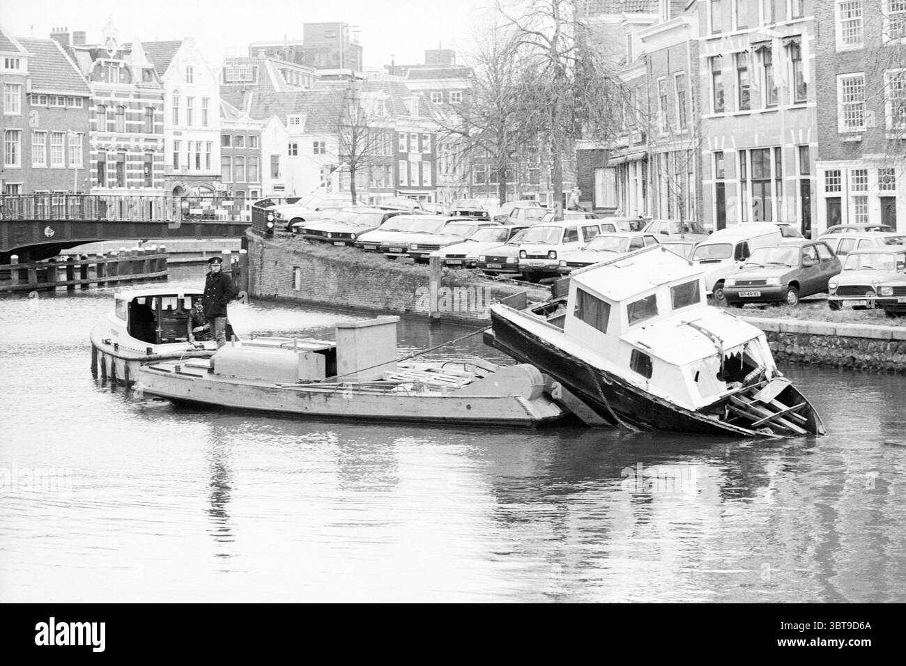 La barca marcio viene trainata da un'altra barca Haarlem Paesi Bassi., Whizgle News, Dutch Desk, Paesi Bassi, 1950 - 2000. Questi sono gli elementi dell'immagine. La scena raffigura un tranquillo canale fiancheggiato da edifici affascinanti caratterizzati da un'architettura tradizionale. Gli edifici presentano un mix di tetti a spiovente e grandi finestre, realizzate in sfumature di grigio e beige, evocando un senso di storia. Una fila di veicoli, principalmente auto d'epoca, riempie la strada lungo il canale, il loro design suggerisce un'epoca passata. In primo piano, due barche riposano sull'acqua ferma. Un'imbarcazione appare più grande Foto Stock