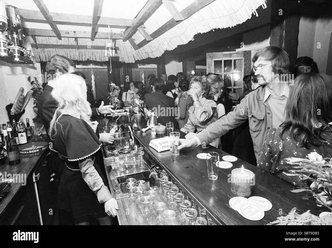 Int Cafe/Bar., Whizgle News, Dutch Desk, Paesi Bassi, 1950 - 2000. Questi sono gli argomenti nell'immagine. La scena raffigura un vivace ambiente bar pieno di persone impegnate nella conversazione e nella socializzazione. L'interno ha un fascino rustico, caratterizzato da travi in legno a vista e da un soffitto basso e accogliente. L'illuminazione è soffusa ma calda, creando un'atmosfera invitante, illuminata da lampade a soffitto che proiettano una luce soffusa in tutta la stanza. Sullo sfondo di bottiglie e bicchieri disposti ordinatamente sugli scaffali del bar, i clienti stanno vicini, creando un senso di cameratismo. Un gruppo di indivi Foto Stock