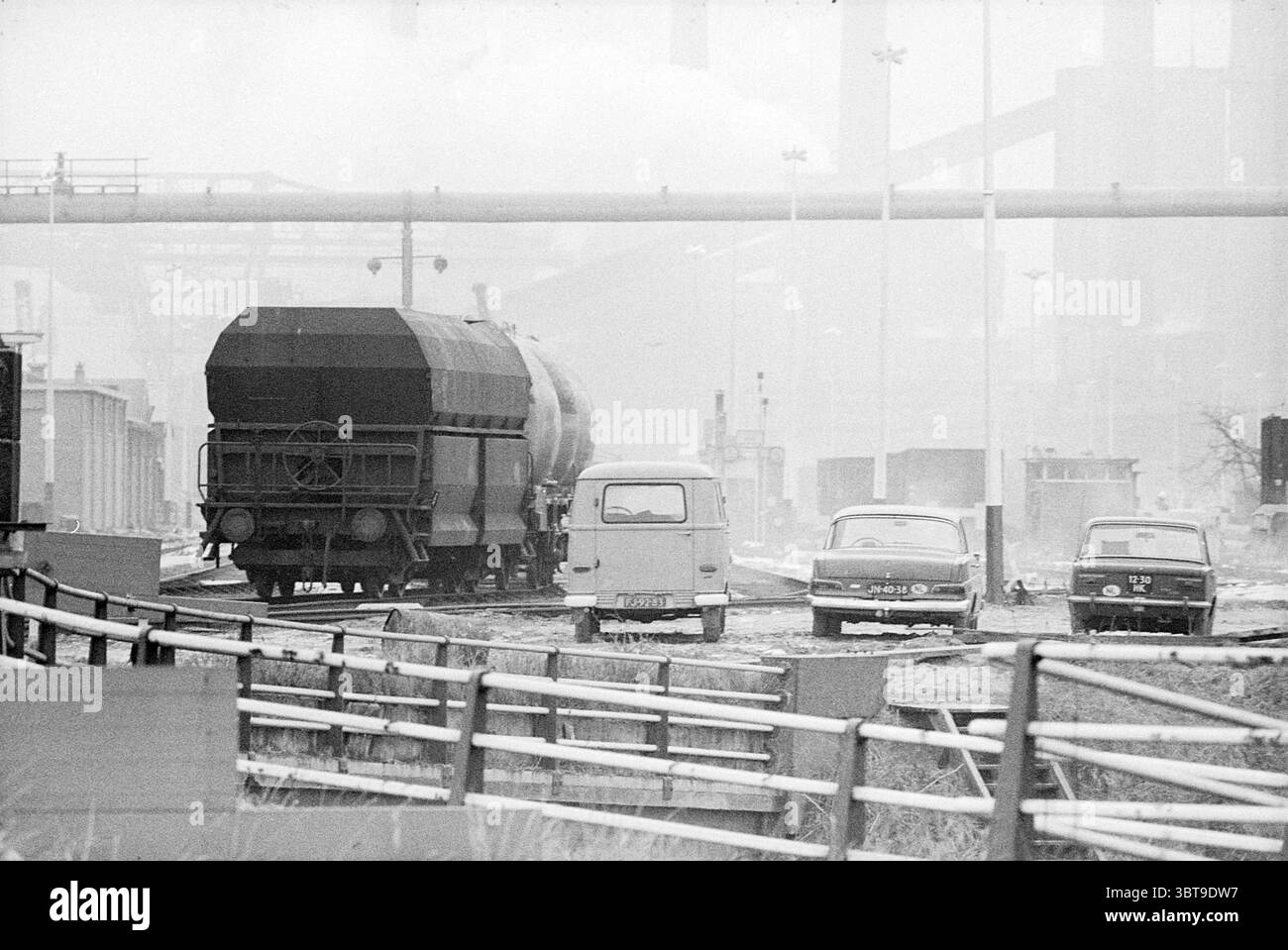 Sciopero ferroviario Hoogovens Hoogovens, Whizgle News, Dutch Desk, Paesi Bassi, 1950 - 2000 il 16-12-1969. Questi sono gli elementi dell'immagine. La scena cattura un paesaggio industriale caratterizzato da un'atmosfera nebbiosa e ricoperta. Un binario ferroviario passa diagonalmente attraverso il centro, portando ad una grande carrozza merci cilindrica che domina il primo piano. Quest'auto, dipinta con un tono tenue, ha una copertura pesante e scura che suggerisce il trasporto di materiali sfusi. Intorno alla linea ferroviaria, è possibile vedere diversi veicoli parcheggiati a caso. Tra questi, un furgone boxy vintage spicca per la sua l Foto Stock
