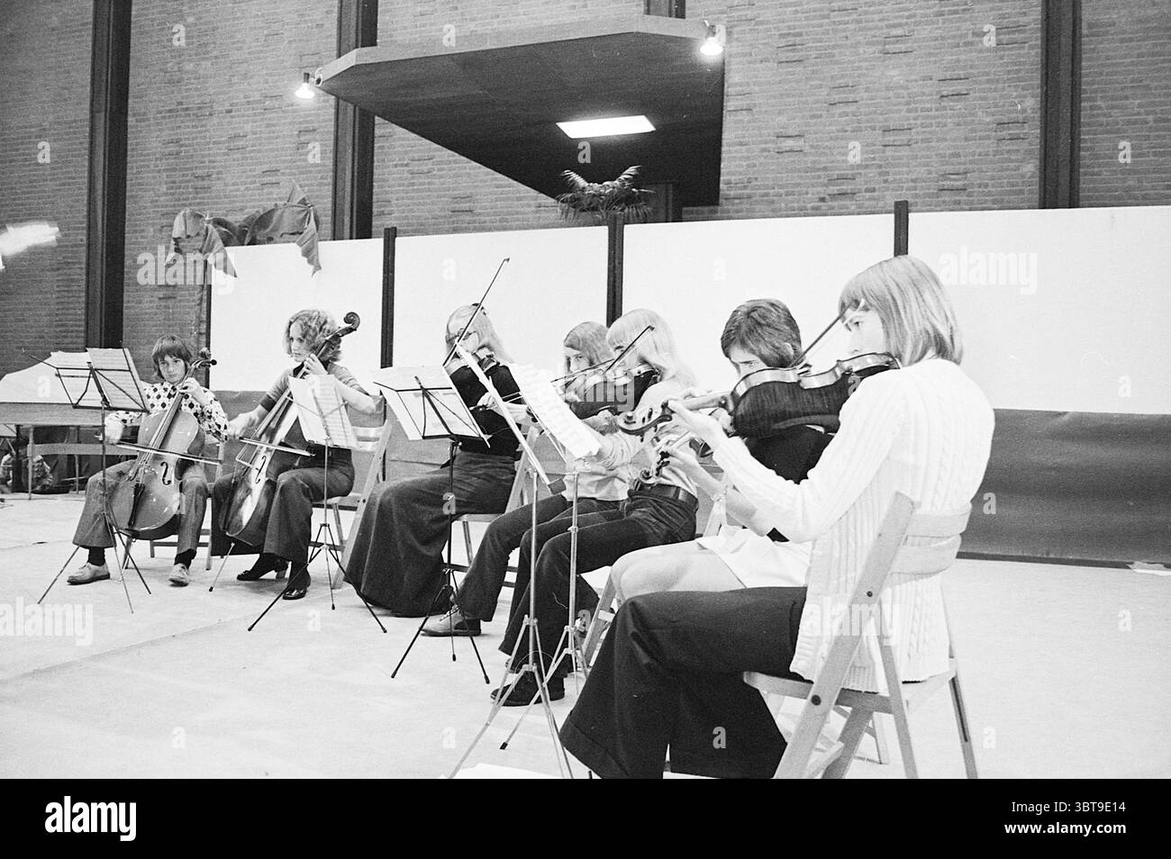 Concert Beverwijk Music School Concerts, Whizgle News, Dutch Desk, Paesi Bassi, 1950 - 2000 il 20-06-1972. Questi argomenti vengono visualizzati nell'immagine. La scena raffigura un gruppo di musicisti impegnati in una performance o prova, disposti in un semicerchio, con particolare attenzione ai loro strumenti e alle dinamiche dell'ensemble. I musicisti includono sei individui: Tre che suonano violini, uno su un violoncello, e gli altri con stili e posture diversi che suggeriscono impegno e concentrazione. In termini di colore, i musicisti indossano un mix di abiti chiari e scuri, combinando motivi e tessuti solidi. In particolare, o Foto Stock