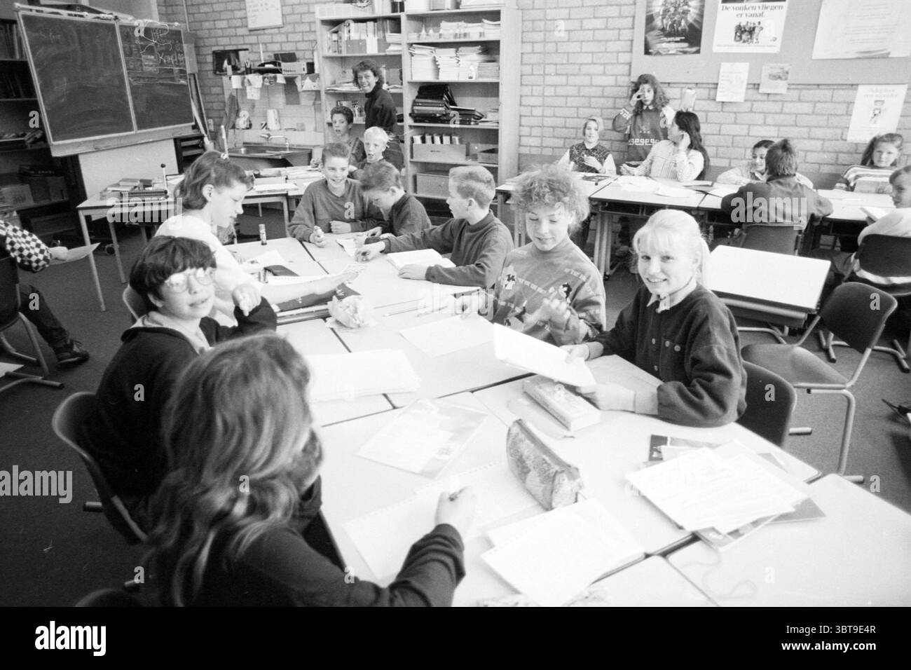 Ragazza belga Miss Ellie in classe Bavoschool Persons Schools Haarlem Paesi Bassi, Whizgle News, Dutch Desk, Paesi Bassi, 1950 - 2000 il 10-04-1990. Questi sono gli argomenti nell'immagine. La scena raffigura un'aula piena di bambini impegnati in varie attività. La composizione è vivace, con alcuni studenti seduti alle scrivanie mentre altri stanno in piedi o interagiscono nelle vicinanze. L'illuminazione è morbida e naturale, probabilmente proveniente da finestre che lasciano entrare la luce del giorno, creando un'atmosfera calda. Le pareti sono adornate con materiali educativi e manifesti, aggiungendo un elemento di colore al resto m Foto Stock