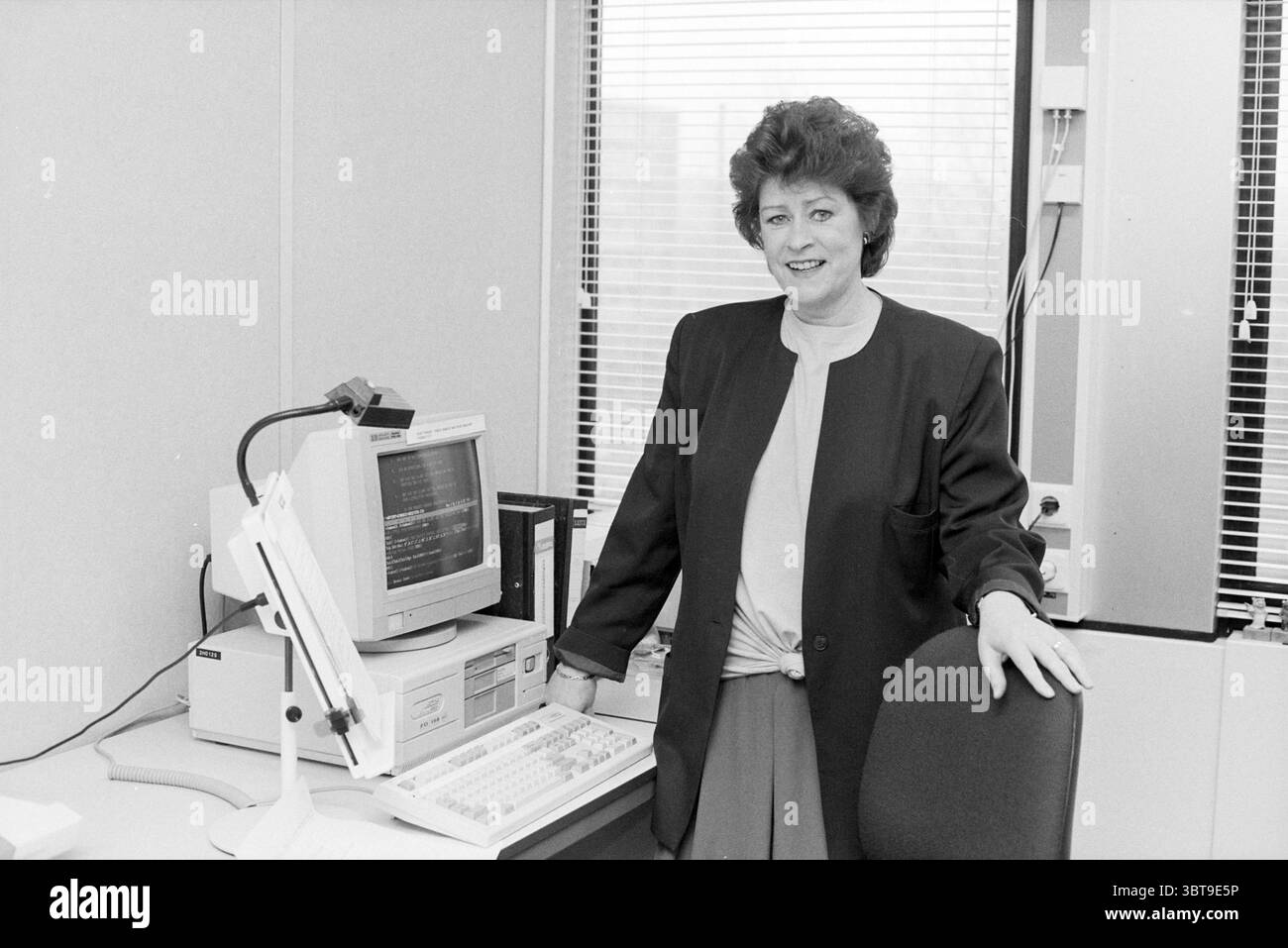 Ritratto di donna con lavagna a fogli mobili e computer, Whizgle News, Dutch Desk, Paesi Bassi, 1950 - 2000 il 12-1991. L'immagine mostra questi argomenti. La scena cattura un ambiente di lavoro professionale con una donna in piedi con sicurezza accanto alla sua scrivania. Ha un aspetto raffinato, caratterizzato da un blazer scuro su una parte superiore di colore chiaro, che crea un contrasto sorprendente. La sua acconciatura è ben curata e si aggiunge al suo comportamento professionale. Lo spazio di lavoro è arredato in modo modesto, con una classica scrivania che include un computer vecchio, abbronzatura e grigio. A sinistra del computer è visibile una tastiera, Foto Stock