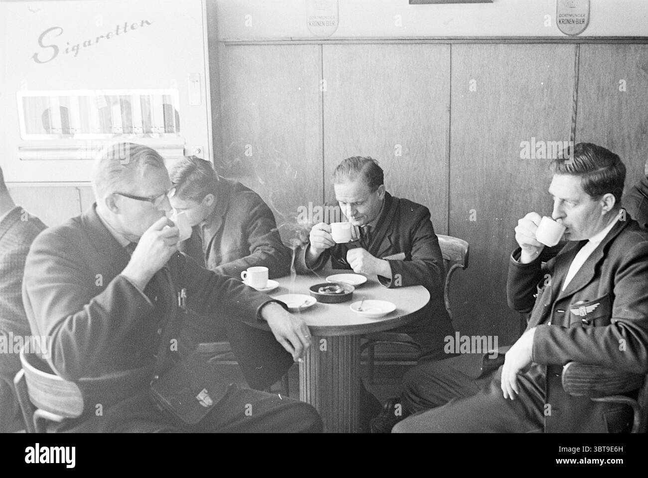 Autisti di caffè Noord Zuid Hollandse Vervoersmaatschappij N.V. NZ, Whizgle News, Dutch Desk, Paesi Bassi, 1950 - 2000 il 20-10-1964. Questi argomenti sono mostrati nell'immagine. La scena presenta un interno accogliente, probabilmente un caffè o un piccolo punto di ritrovo. Ci sono diversi uomini seduti intorno a un tavolo rotondo di legno, ciascuno con una piccola tazza bianca in mano, sorseggiando da esso mentre si impegnano in una conversazione tranquilla. Gli uomini esibiscono una gamma di abbigliamento, per lo più abiti scuri, dando un senso di formalità mescolato a un'atmosfera informale. Un uomo, con gli occhiali, siede prominente a sinistra, profondamente concentrato sul suo dr Foto Stock
