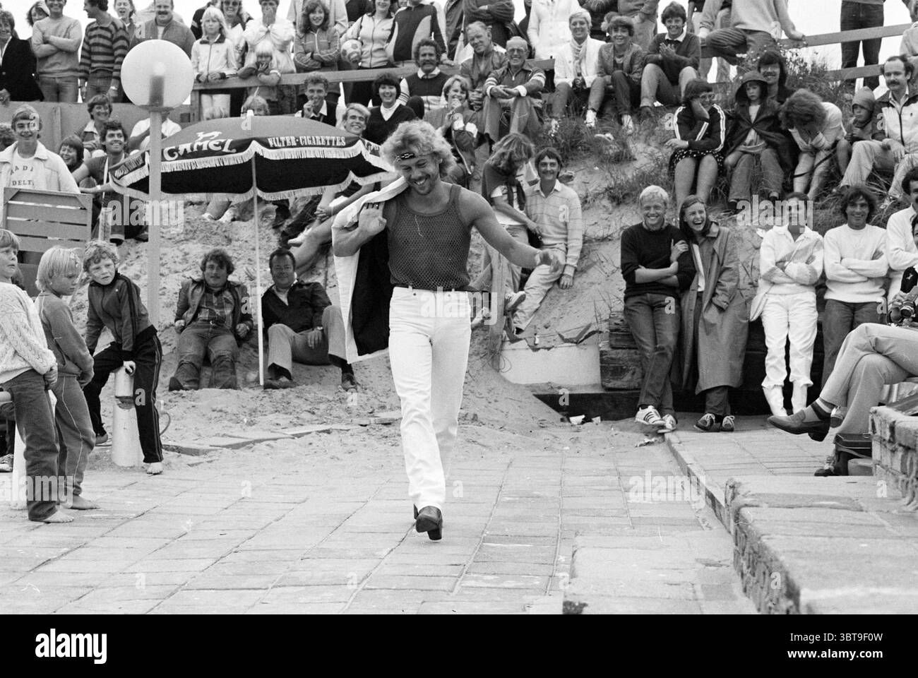 Miss Beach Zandvoort IV Miss concorso Zandvoort Zandvoort Boulevard, Whizgle News, Dutch Desk, Paesi Bassi, 1950 - 2000 il 08-08-1981. Questi argomenti vengono visualizzati nell'immagine. La scena cattura un vivace evento all'aperto, probabilmente in una spiaggia o in una località balneare, con un grande pubblico riunito in una disposizione a più livelli su un terreno sabbioso. La folla è variegata, con persone di varie età e stili, tutte coinvolte nell'azione in primo piano. Al centro, un uomo con un comportamento sicuro cammina con un sorriso gioviale, vestito con un top a rete aderente e pantaloni bianchi. La sua postura trasuda cha Foto Stock