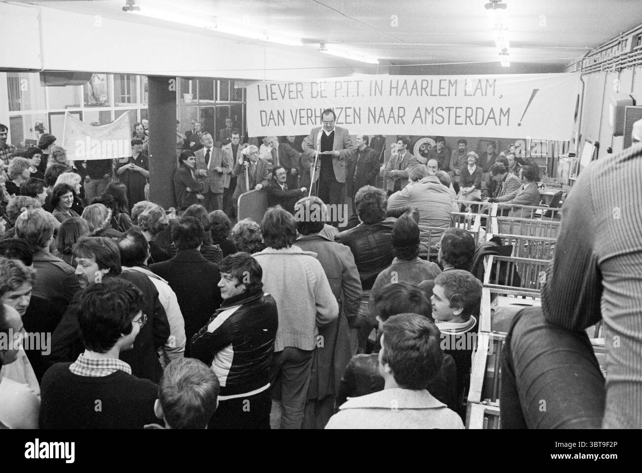 Riunione dimostrativa PTT Bailjuwslaan dimostrazione PTT servizi postali città post Haarlem Baljuwslaan Paesi Bassi, Whizgle News, Dutch Desk, Paesi Bassi, 1950 - 2000 il 09-01-1980. Questi argomenti sono mostrati nell'immagine. La scena è piena di energia, dominata da una grande folla riunita in un interno spazioso, forse un municipio o un ufficio. Lo spazio è ben illuminato, con luci luminose che proiettano un bagliore caldo ma leggermente ruvido sopra la raccolta. In primo piano, un gruppo eterogeneo di persone è raggruppato intorno a una figura centrale che si trova su un podio. L'altoparlante, vestito Foto Stock