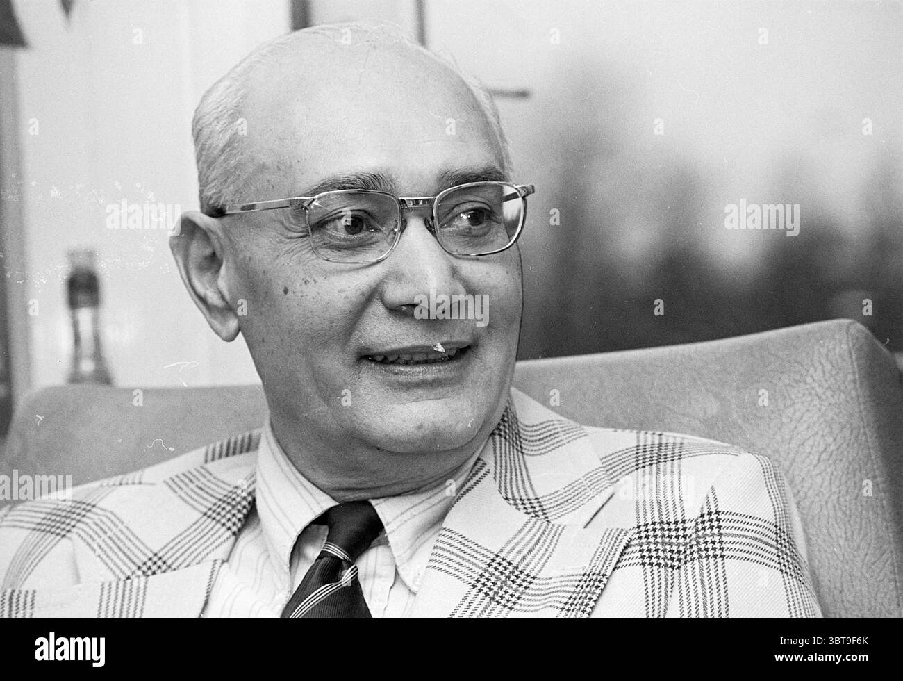 Ritratto di un gentiluomo calvo con occhiali e una giacca a scacchi, Whizgle News, Dutch Desk, Paesi Bassi, 1950 - 2000. Questi argomenti sono mostrati nell'immagine. Un gentiluomo siede comodamente in un ambiente ben arredato, caratterizzato da una lussuosa poltrona di colore chiaro. Ha un sorriso leggero e affabile che trasmette calore e avvicinabilità. Il suo abbigliamento è costituito da una giacca a quadri dalle tonalità morbide del bianco e del grigio, abbinata a una camicia bianca formale e a una cravatta scura che aggiunge contrasto al suo outfit. Lo sfondo presenta un ambiente neutro e leggermente sfocato, che suggerisce uno spazio interno accogliente, perha Foto Stock