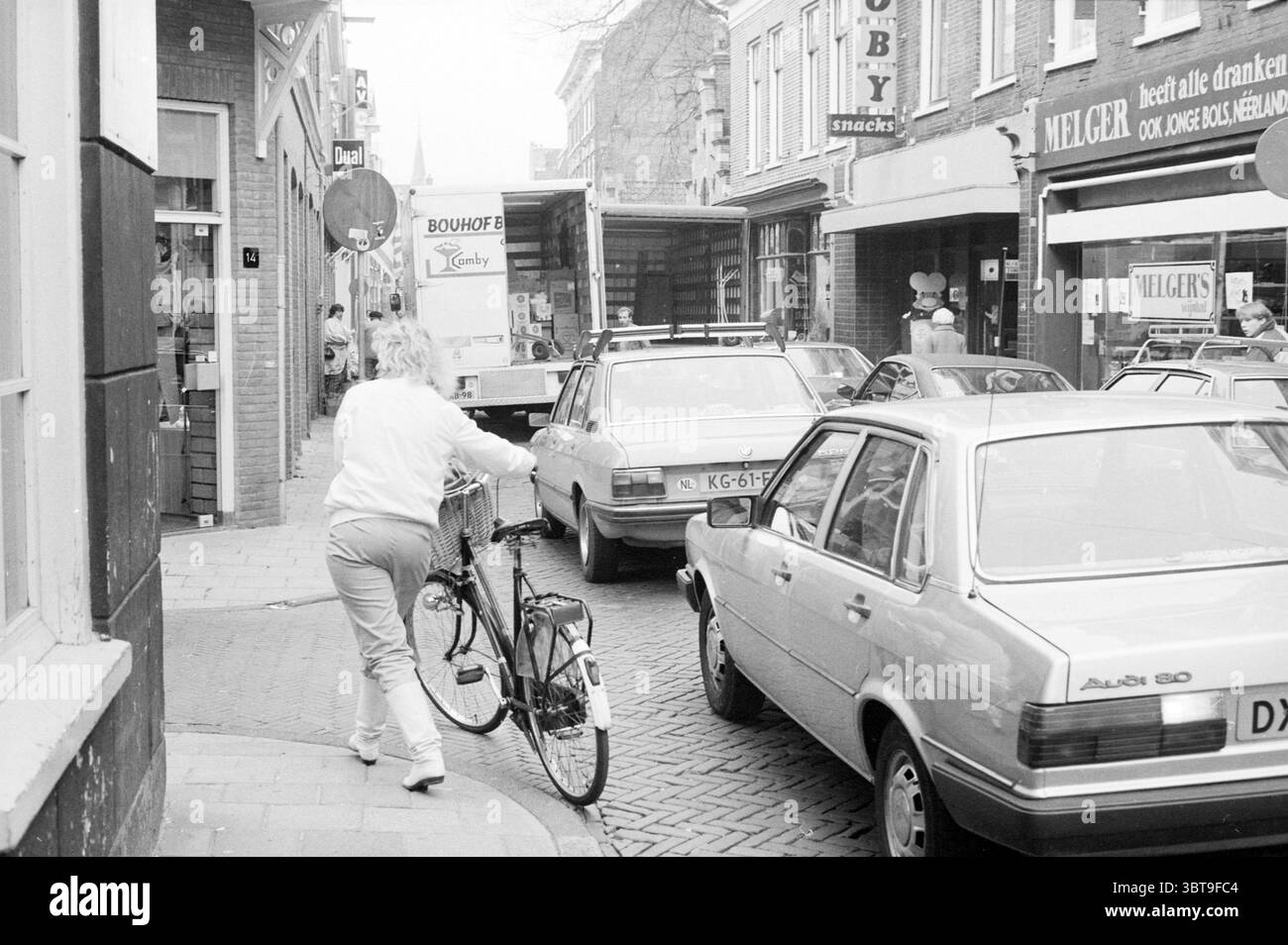 Problemi di traffico Barrevoetsstraat traffico sicuro Haarlem Barrevoetestraat Paesi Bassi, Whizgle News, Dutch Desk, Paesi Bassi, 1950 - 2000 il 18-04-1984. Questi argomenti vengono visualizzati nell'immagine. La scena cattura una vivace strada urbana piena di veicoli e persone. Verso il primo piano, si vede una persona con una tuta bianca che spinge una bicicletta lungo il marciapiede, suggerendo un ambiente informale e quotidiano. I capelli biondi della persona si distinguono contro i toni tenui della strada, contribuendo a creare un'atmosfera vivace. Numerose auto fiancheggiano la strada, dai modelli vintage t Foto Stock