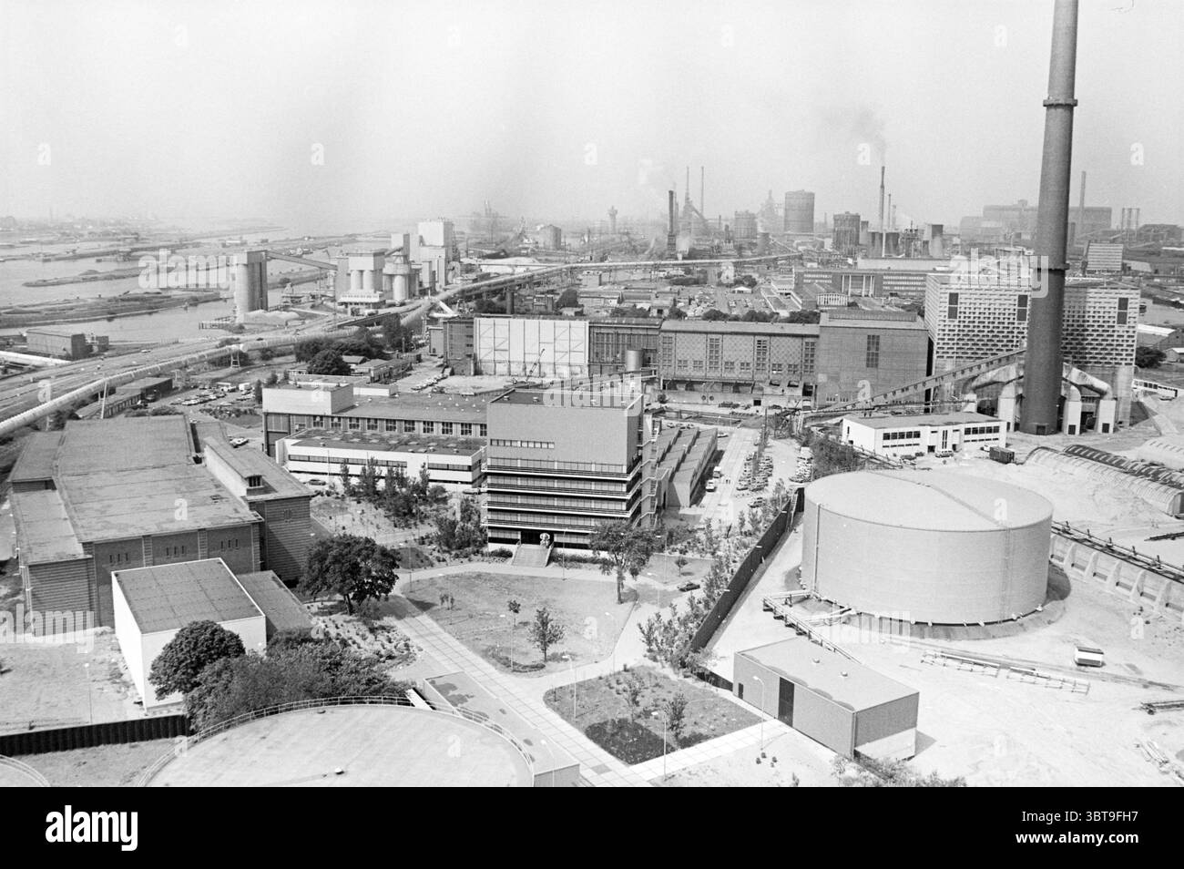 Canale del Mare del Nord e dintorni con industria e case Velsen, Whizgle News, Dutch Desk, Paesi Bassi, 1950 - 2000 il 02-06-1982. Questi argomenti vengono visualizzati nell'immagine. La scena presenta un vasto paesaggio industriale, caratterizzato da varie strutture ed elementi che evocano un senso di sviluppo urbano. Grandi e funzionali edifici dominano il primo piano, caratterizzati da design geometrici con una miscela di esterni in cemento e acciaio. Le loro tonalità grigie e silenziose contribuiscono a creare un ambiente complessivamente austero, riflettendo un'estetica utilitaria. Al centro, i serbatoi cilindrici più grandi si innalzano Foto Stock